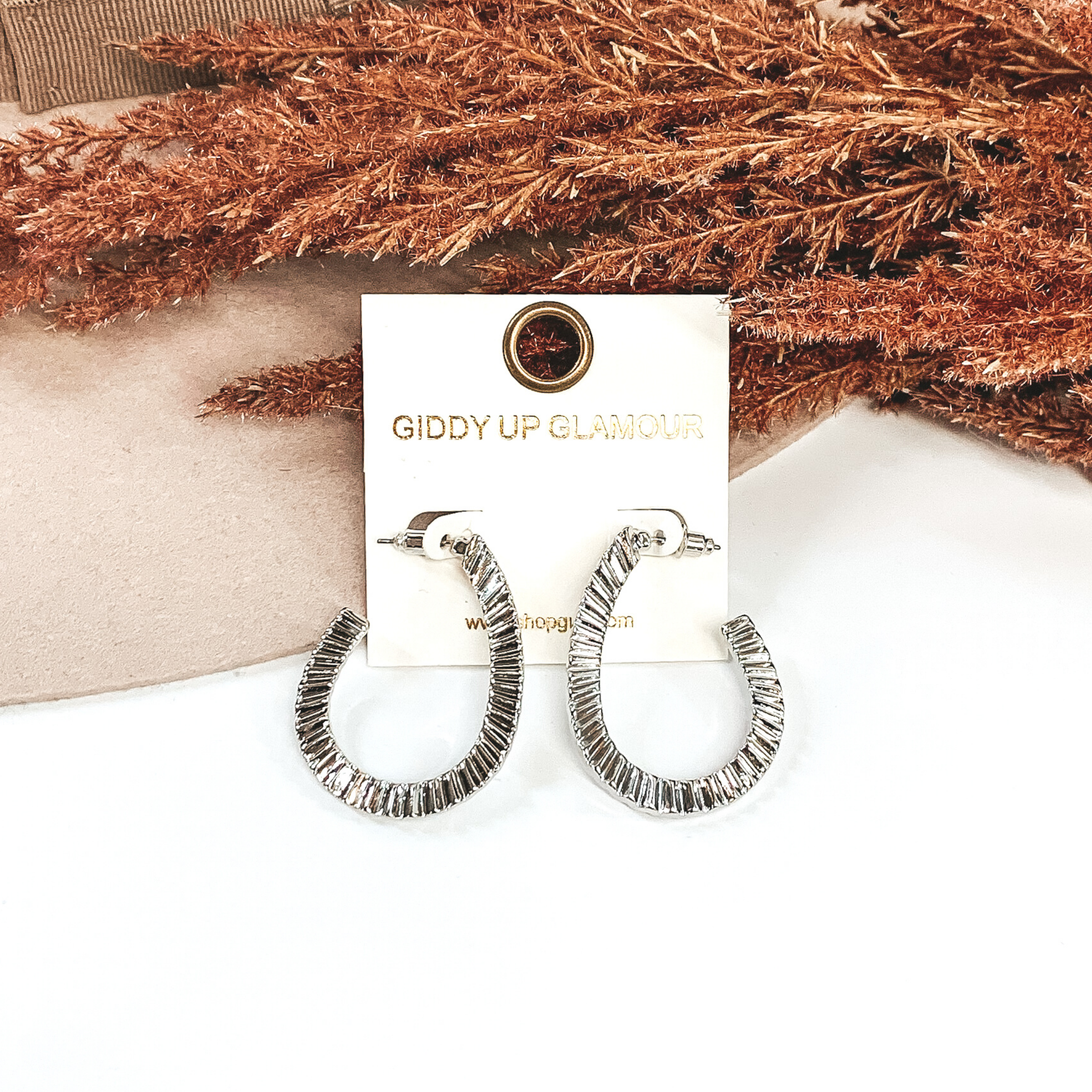 Textured open teardrop earrings in silver that are pictured hanging on a white square earring holder. These earrings are laid on a white and beige background with some brown floral at the top.