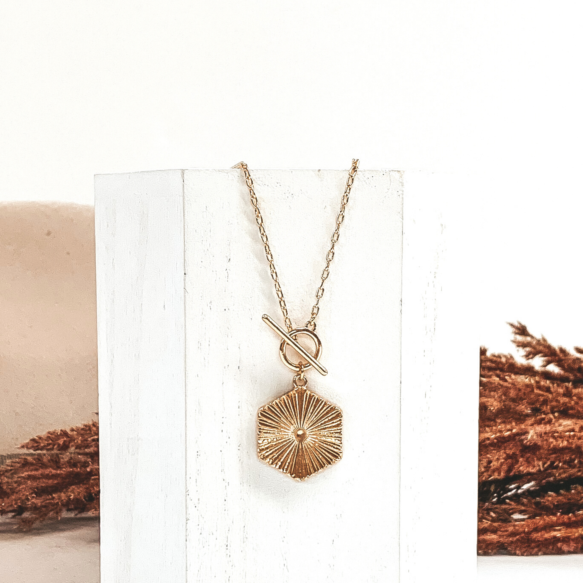 Gold chain necklace with toggle clasp and a hanging gold hexagon pendant. This necklace is laying on a piece of white wood that is in front of a white and beige background with brown floral. 