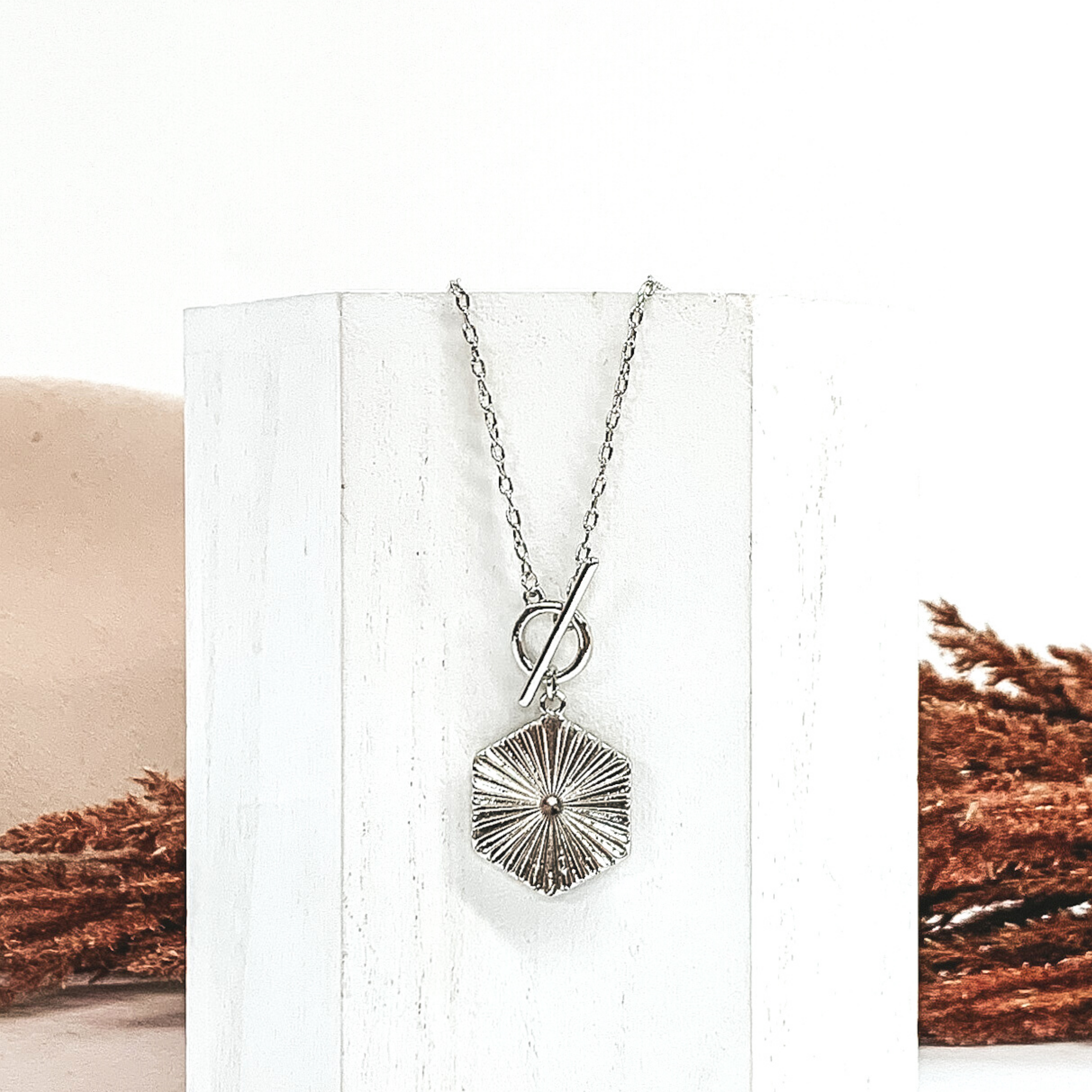 Silver chain necklace with toggle clasp and a hanging silver hexagon pendant. This necklace is laying on a piece of white wood that is in front of a white and beige background with brown floral.