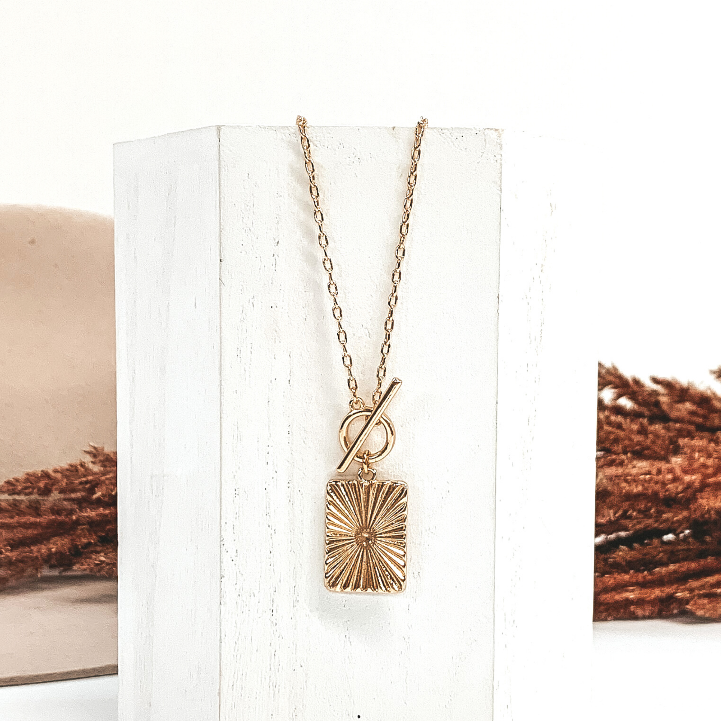 Gold chain necklace with toggle clasp and a hanging gold rectangle pendant. This necklace is laying on a piece of white wood that is in front of a white and beige background with brown floral.