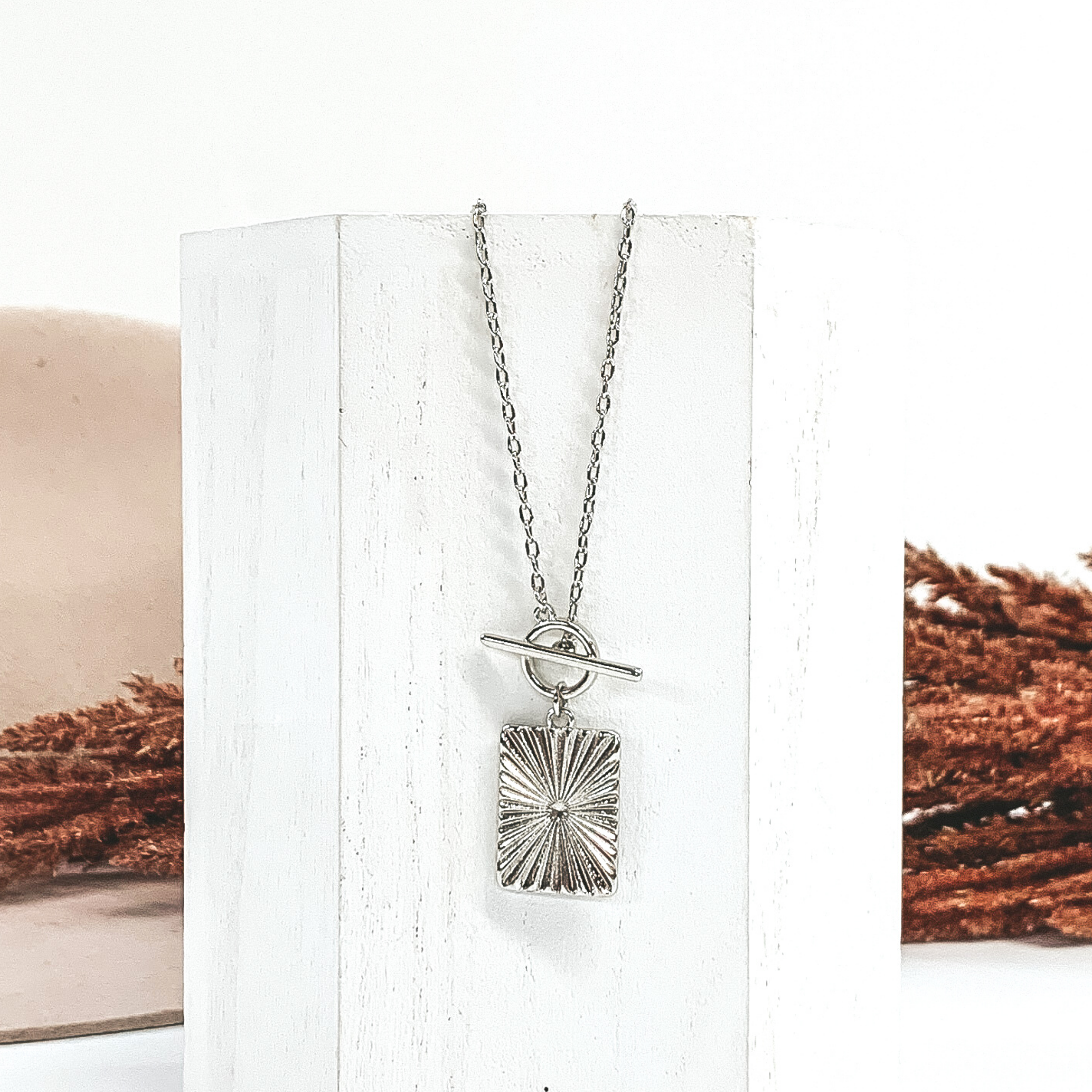 Silver chain necklace with toggle clasp and a hanging silver rectangle pendant. This necklace is laying on a piece of white wood that is in front of a white and beige background with brown floral.