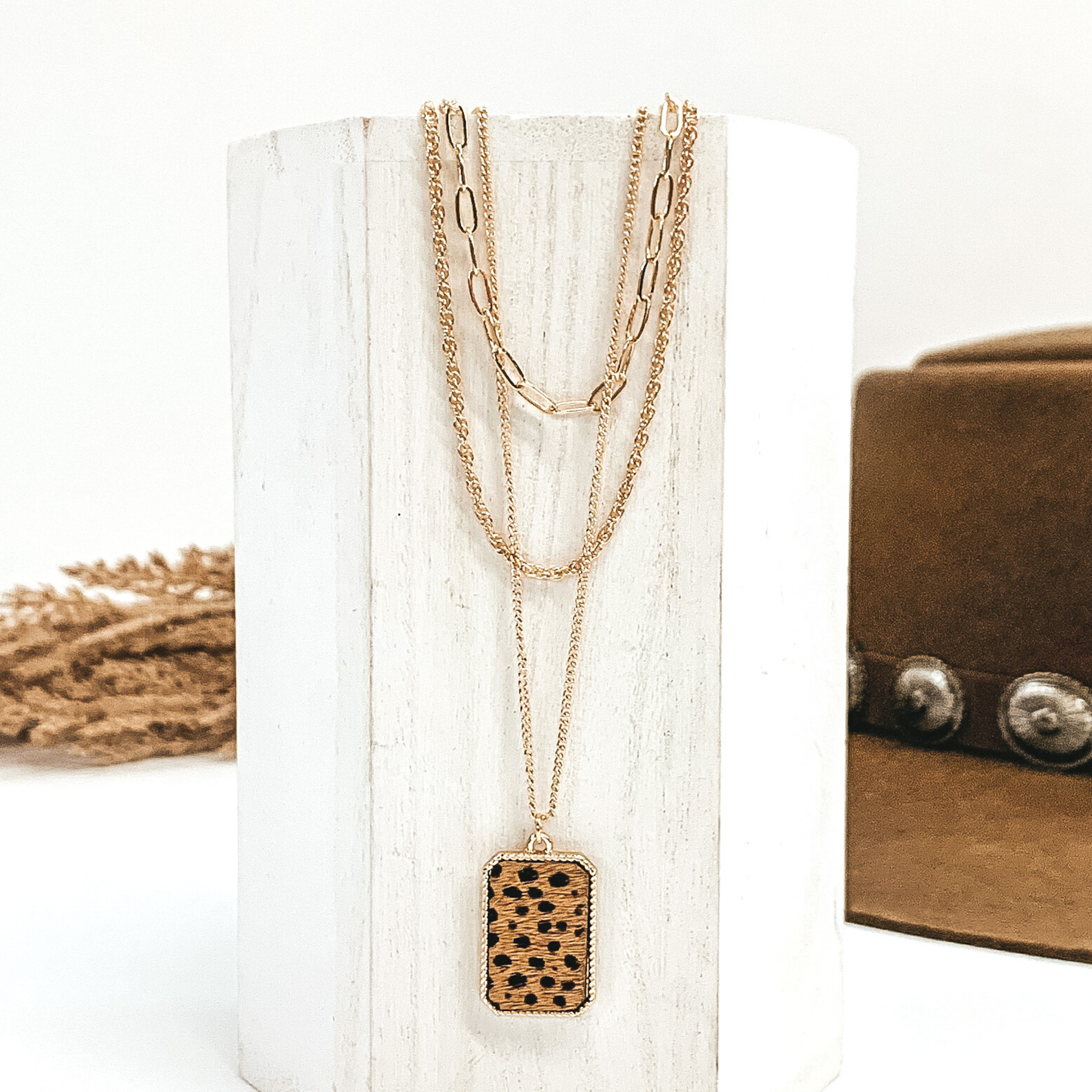 There are three different gold chains at different lengths laying on a white piece of wood. The longest chain has gold rectangle pendant with a brown and black dotted print inlay. This is all pictured on a white background with a brown hat and brown floral in the background.