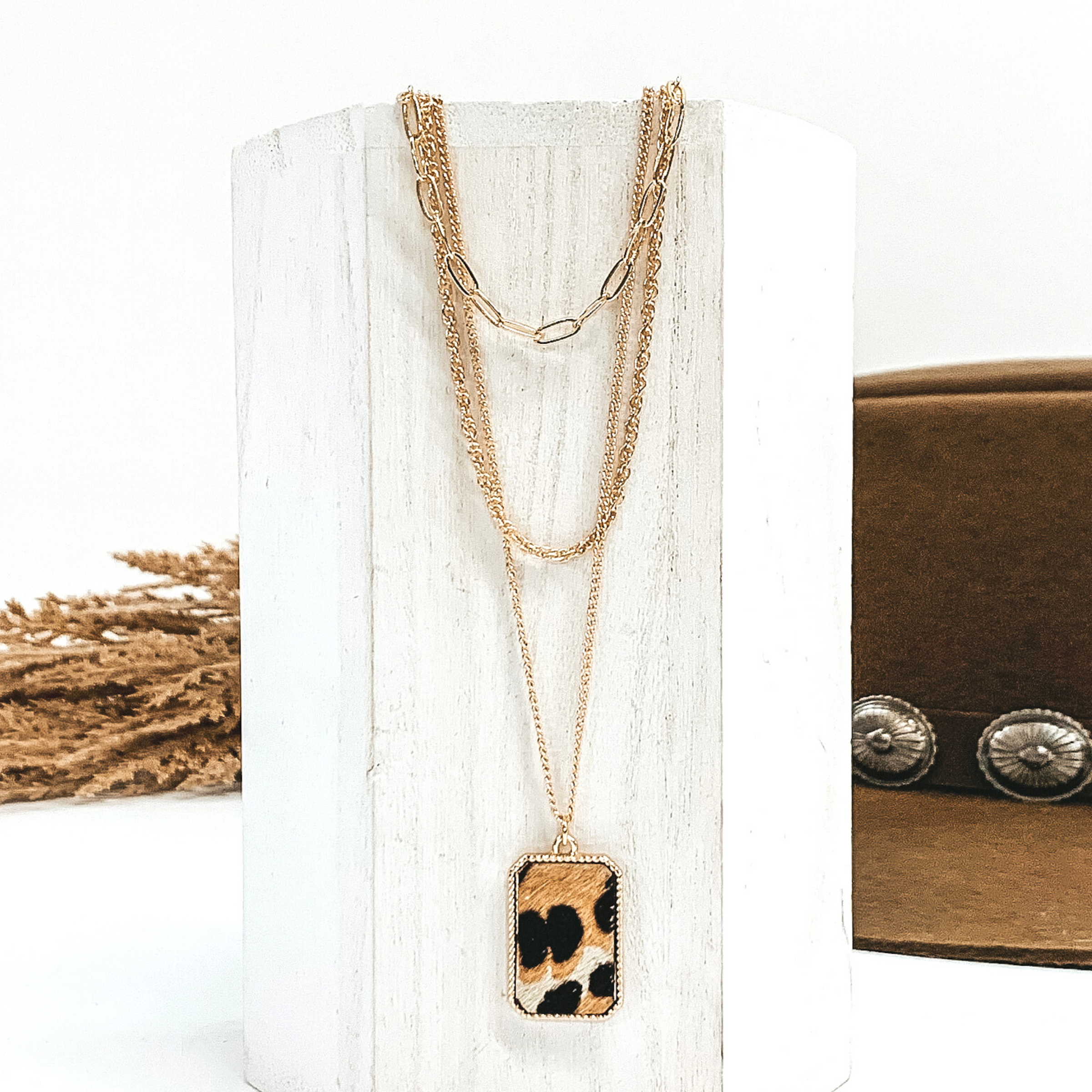 There are three different gold chains at different lengths laying on a white piece of wood. The longest chain has gold rectangle pendant with a ivory, tan, and black animal print inlay. This is all pictured on a white background with a brown hat and brown floral in the background.