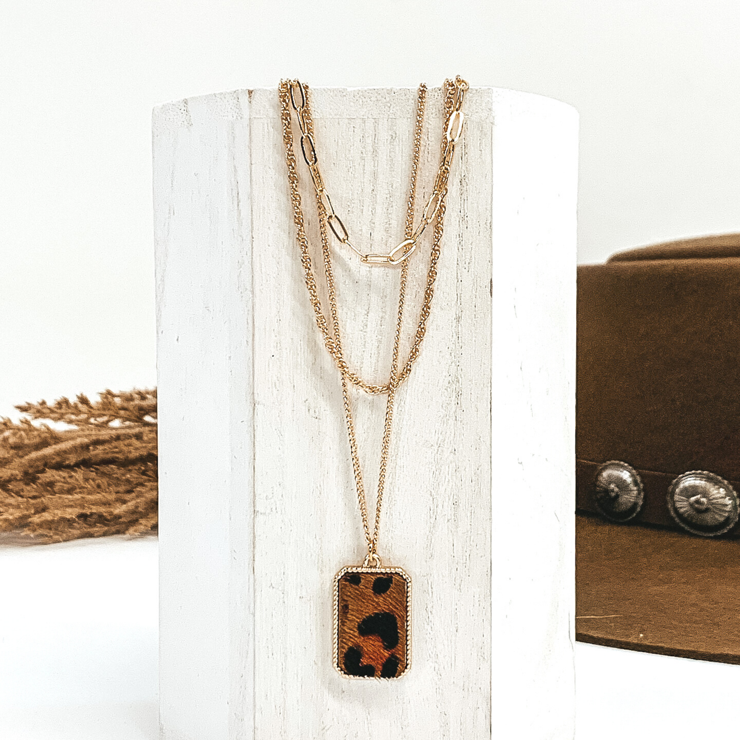 There are three different gold chains at different lengths laying on a white piece of wood. The longest chain has gold rectangle pendant with a brown and black animal print inlay. This is all pictured on a white background with a brown hat and brown floral in the background.