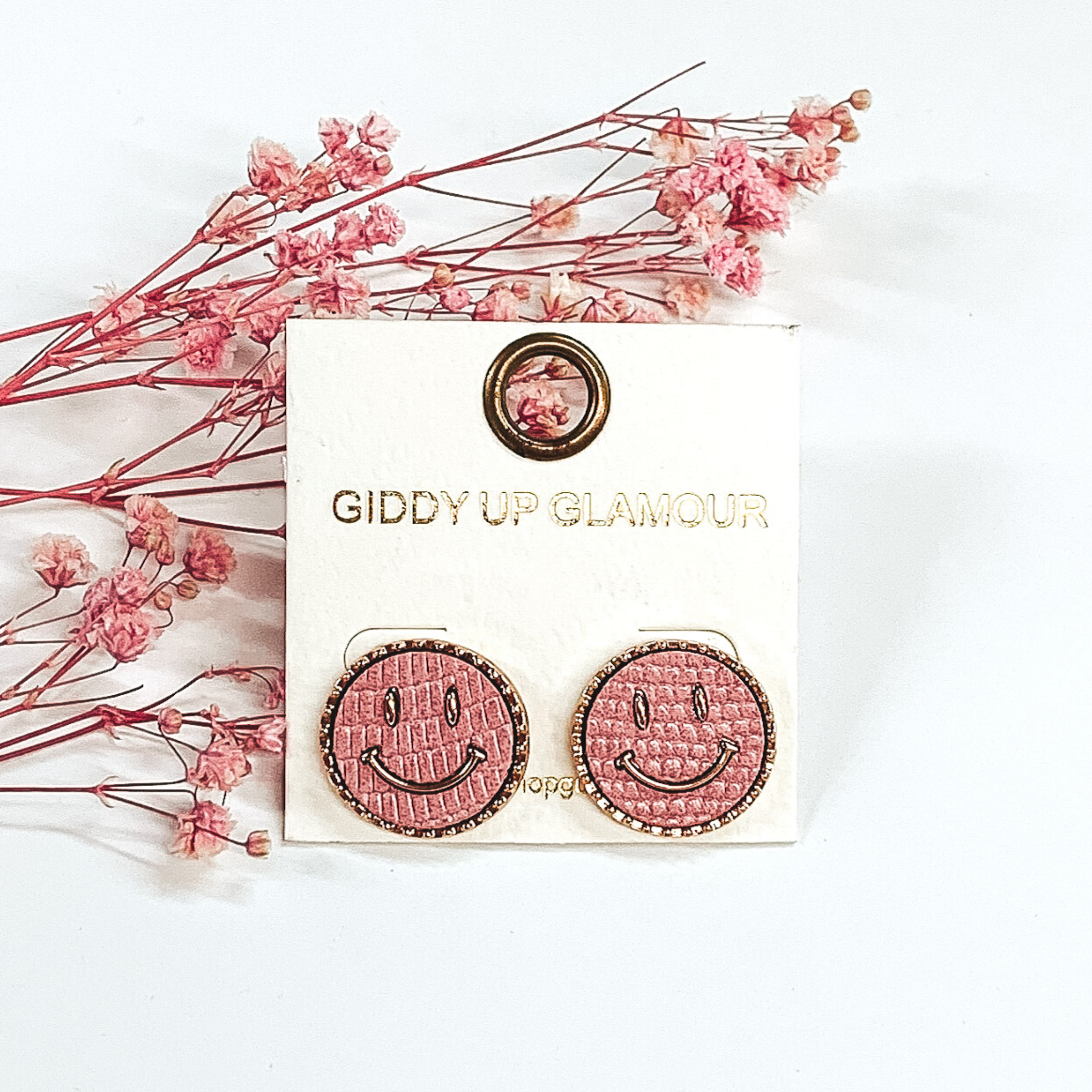 Gold round stud earrings with a pink animal print inlay and a gold smiley face. These earrings are pictured a white background with pink babys breath.