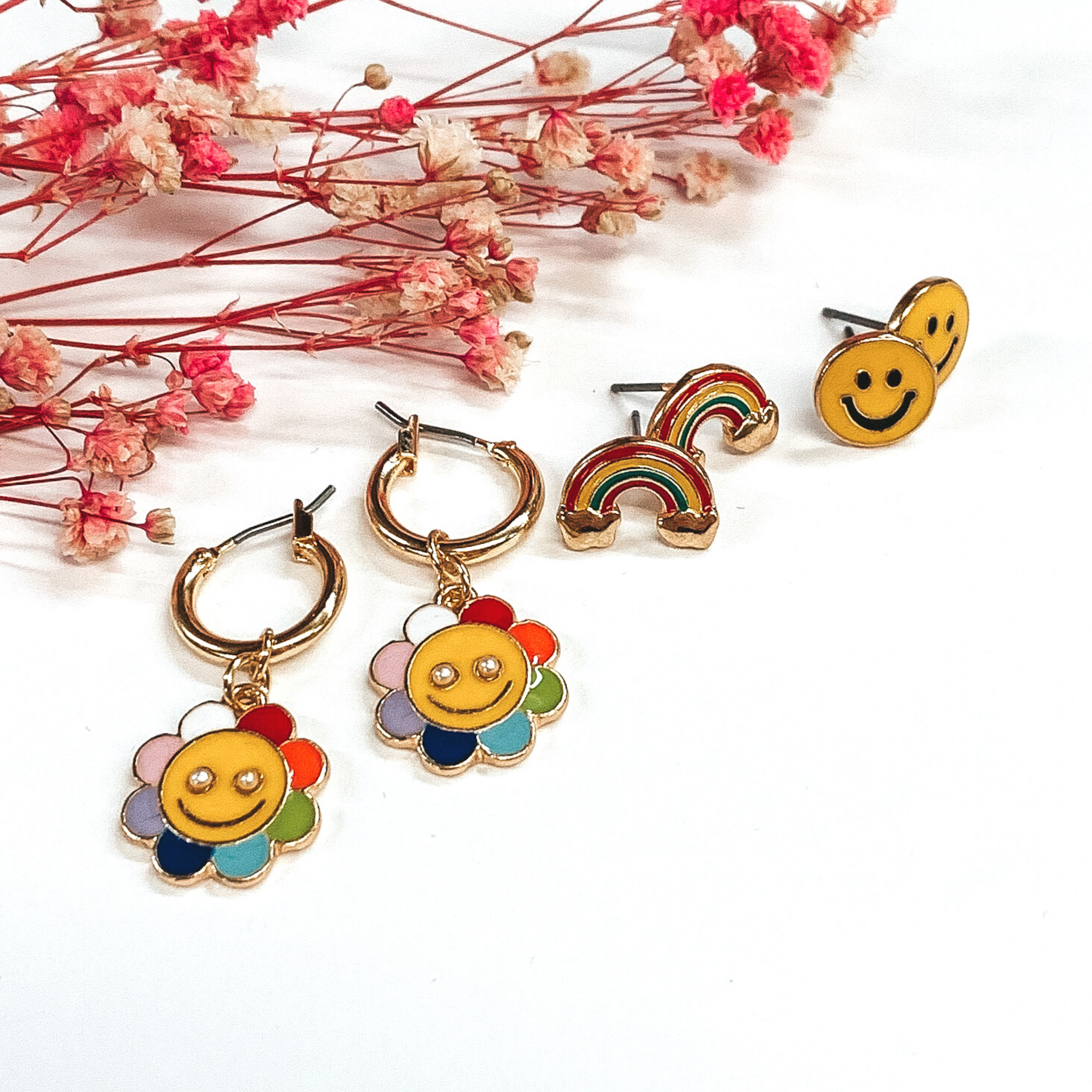 This earring set includes yellow smiley face studs, rainbow studs, and tiny gold hoops with multicolored flower pendants with a smiley face in the center. These earrings are pictured on a white background with pink babys breath. 
