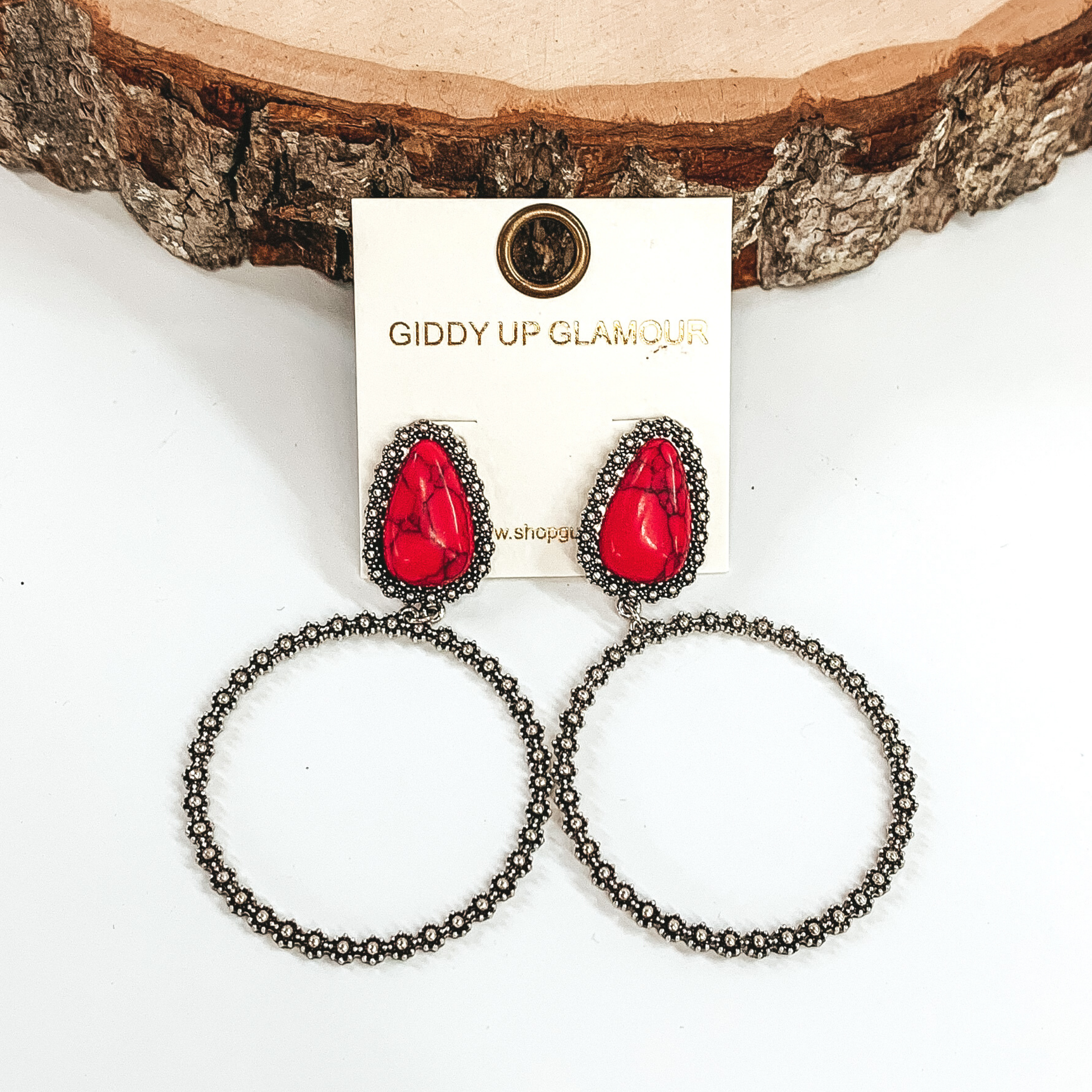 These earrings have red teardrop stone studs outlined in silver. At the bottom of the stones there is a hanging silver circle. These earrings are pictured on a white background laying against a piece of wood.