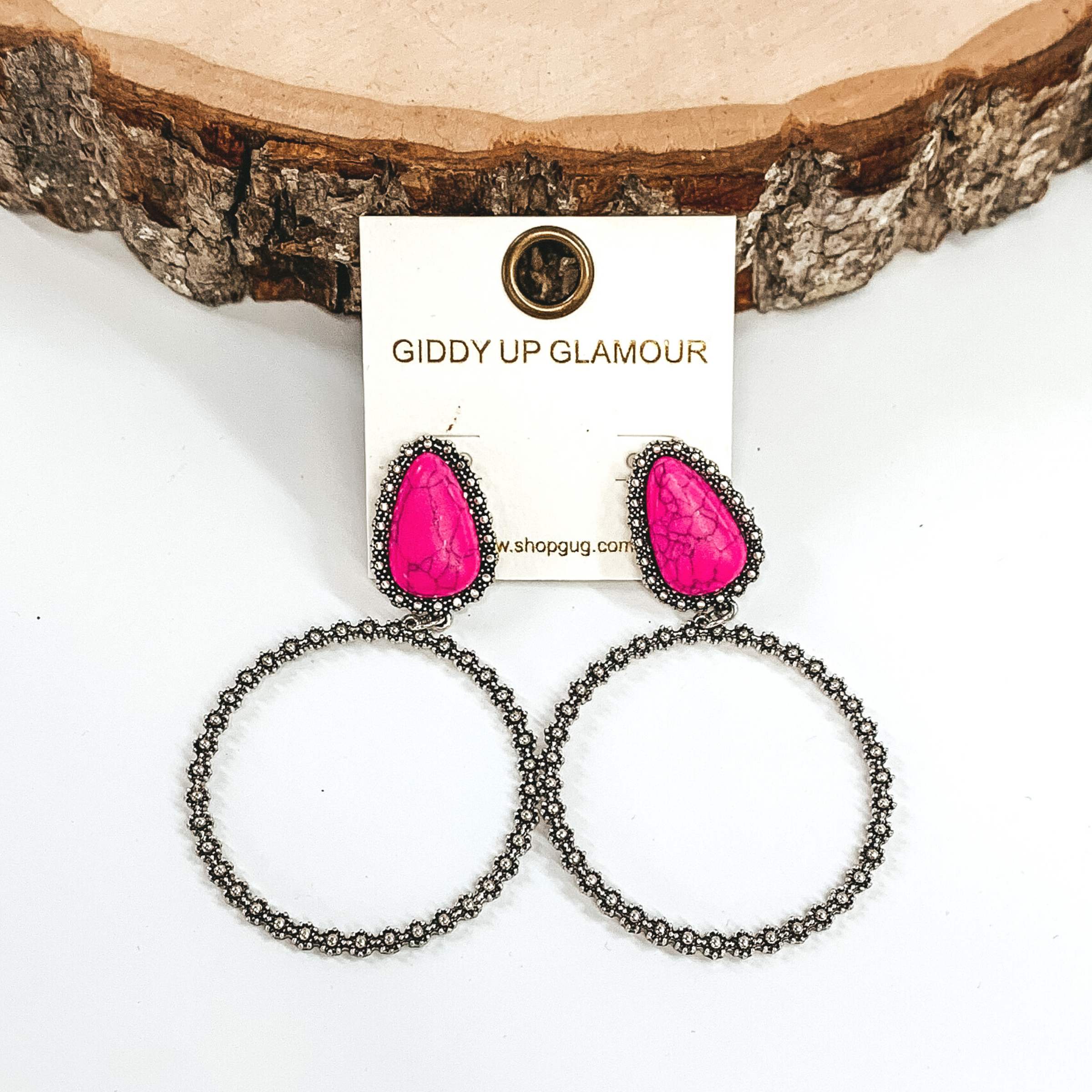 These earrings have pink teardrop stone studs outlined in silver. At the bottom of the stones there is a hanging silver circle. These earrings are pictured on a white background laying against a piece of wood. 