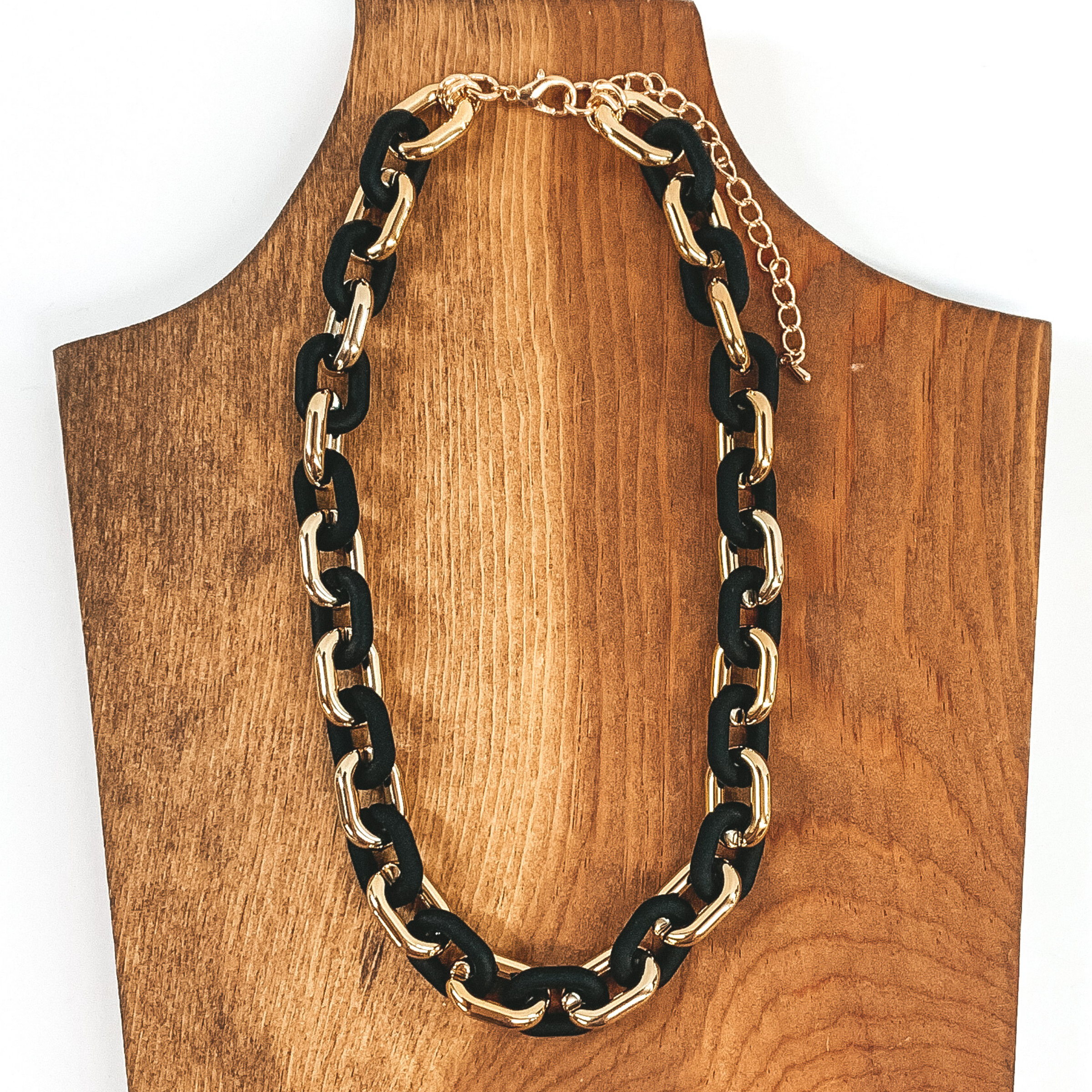 This necklace has thick gold and matte black links that repeat. It has a gold adjustable chain. This necklace is pictured of a piece of wood that is on a white background. 