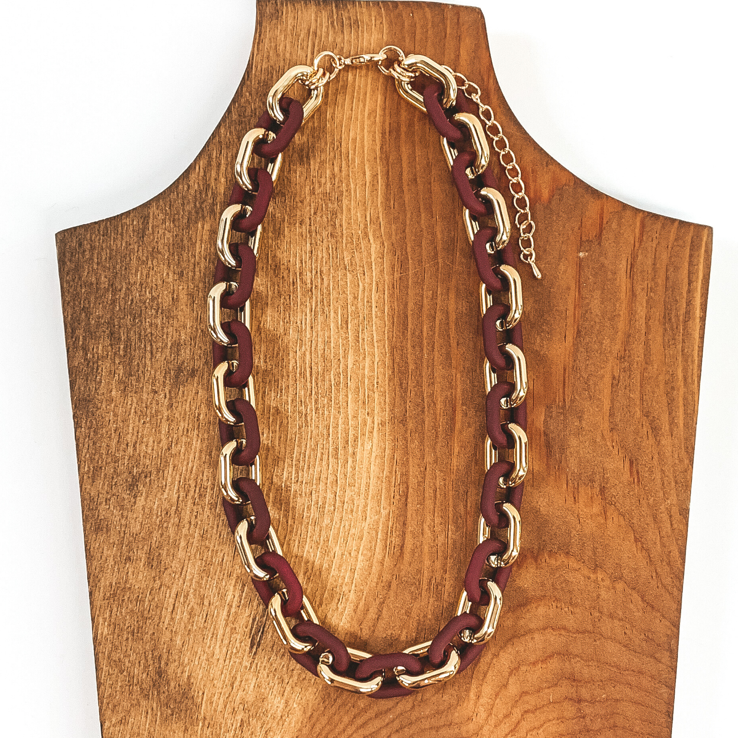 This necklace has thick gold and matte maroon links that repeat. It has a gold adjustable chain. This necklace is pictured of a piece of wood that is on a white background.