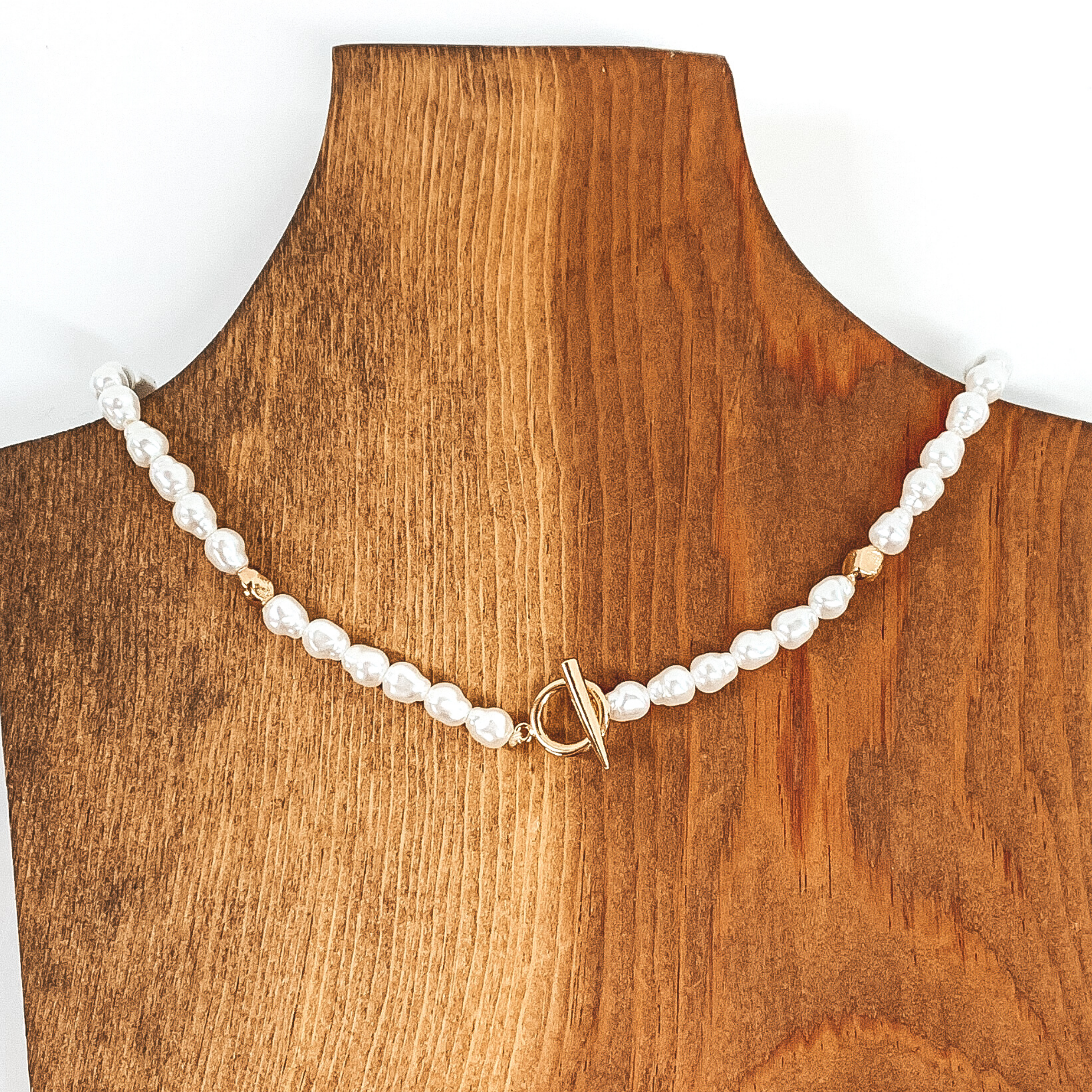 This necklace has white pearl like beads with one gold bead on each side of the gold toggle clasp. This necklace is pictured laying on a piece of wood colored necklace holder on a white background. 