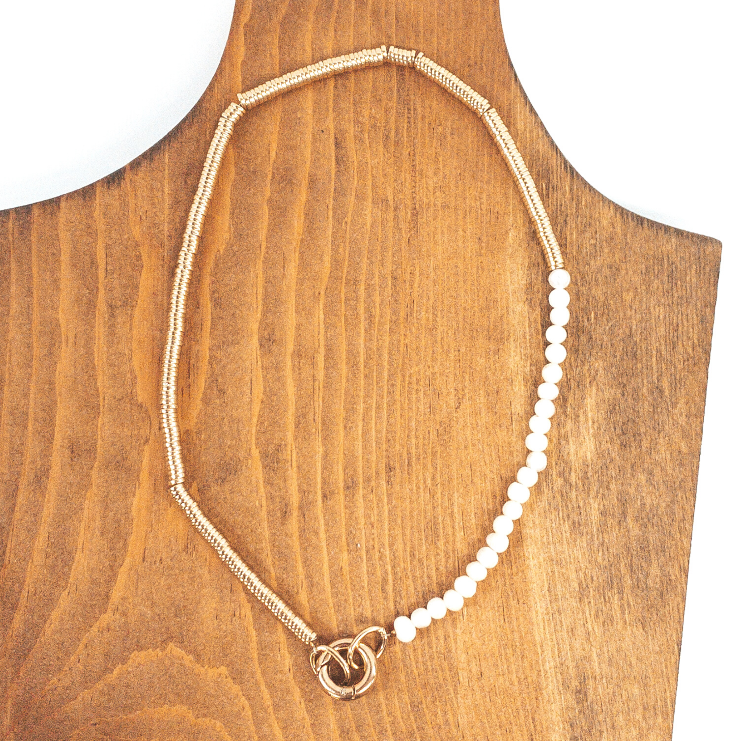 Three quarters of the necklace is gold metal disk beads, while the last quarter is white pearl like beads. There is a gold clasp to close it. This necklace is pictured on a wood necklace holder on a white background. 