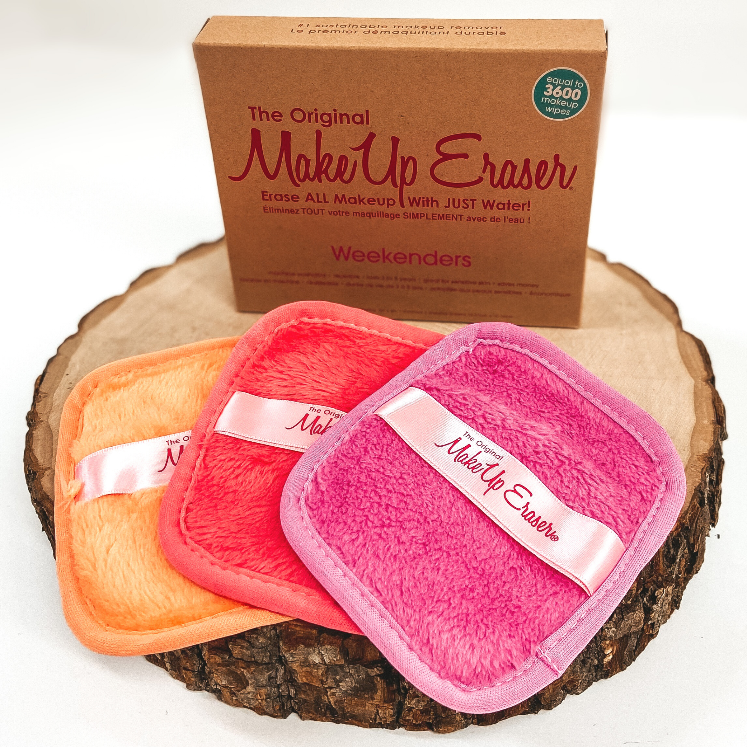 There are three small, rounded rectangle cloths laying on top of each other with light pink tags across the middle of each cloths. These clothes are laying on a piece of wood with the pink cloth laying on top of the coral cloth that is laying on an orange cloth. Behind these cloths is a brown box standing up right on the piece of wood. This is all pictured on a white background.