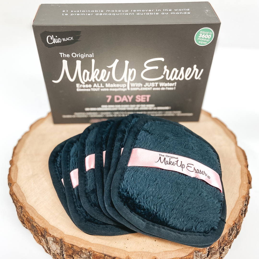 There are seven black, rounded rectangle cloths with a pink tag going through the middle of it and they are laying on top of each other. These cloths are laying on a piece of wood with a black box standing up right behind the cloths. All of this is pictured on a white background.