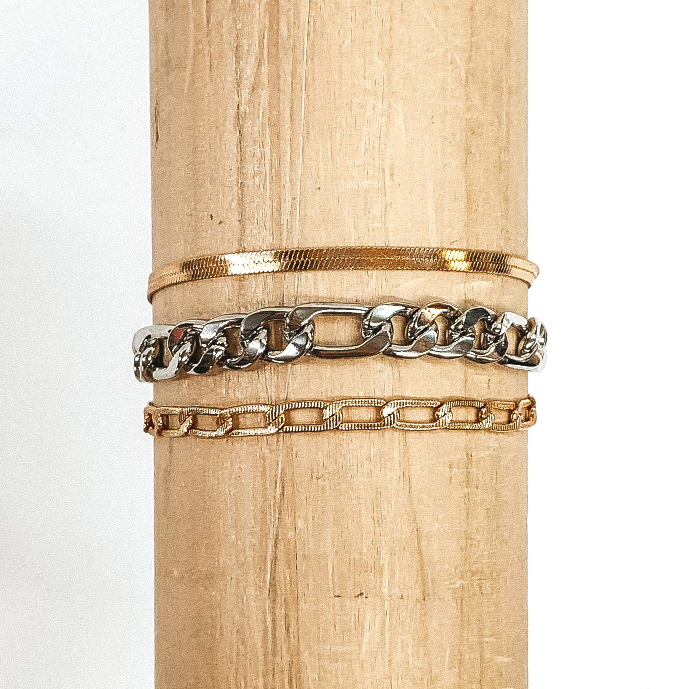 This bracelet includes a gold snake chain, a silver figaro chain, and a gold classic chain.. This bracelet is wrapped around a piece of wood that is pictured on a white background.