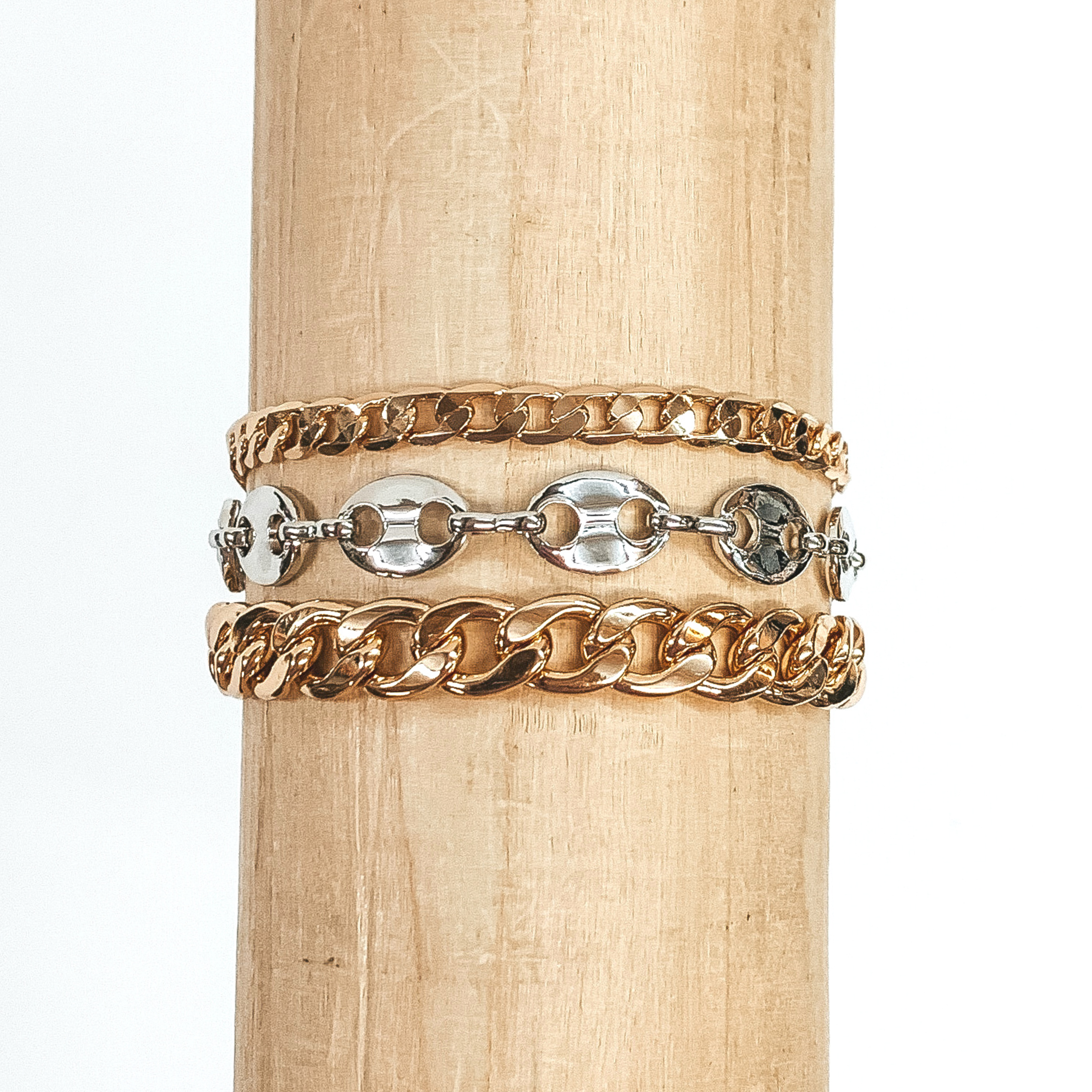 This bracelet includes a flat curb chain, a silver anchor chain, and a gold curb chain. This bracelet is wrapped around a piece of wood that is pictured on a white background. 