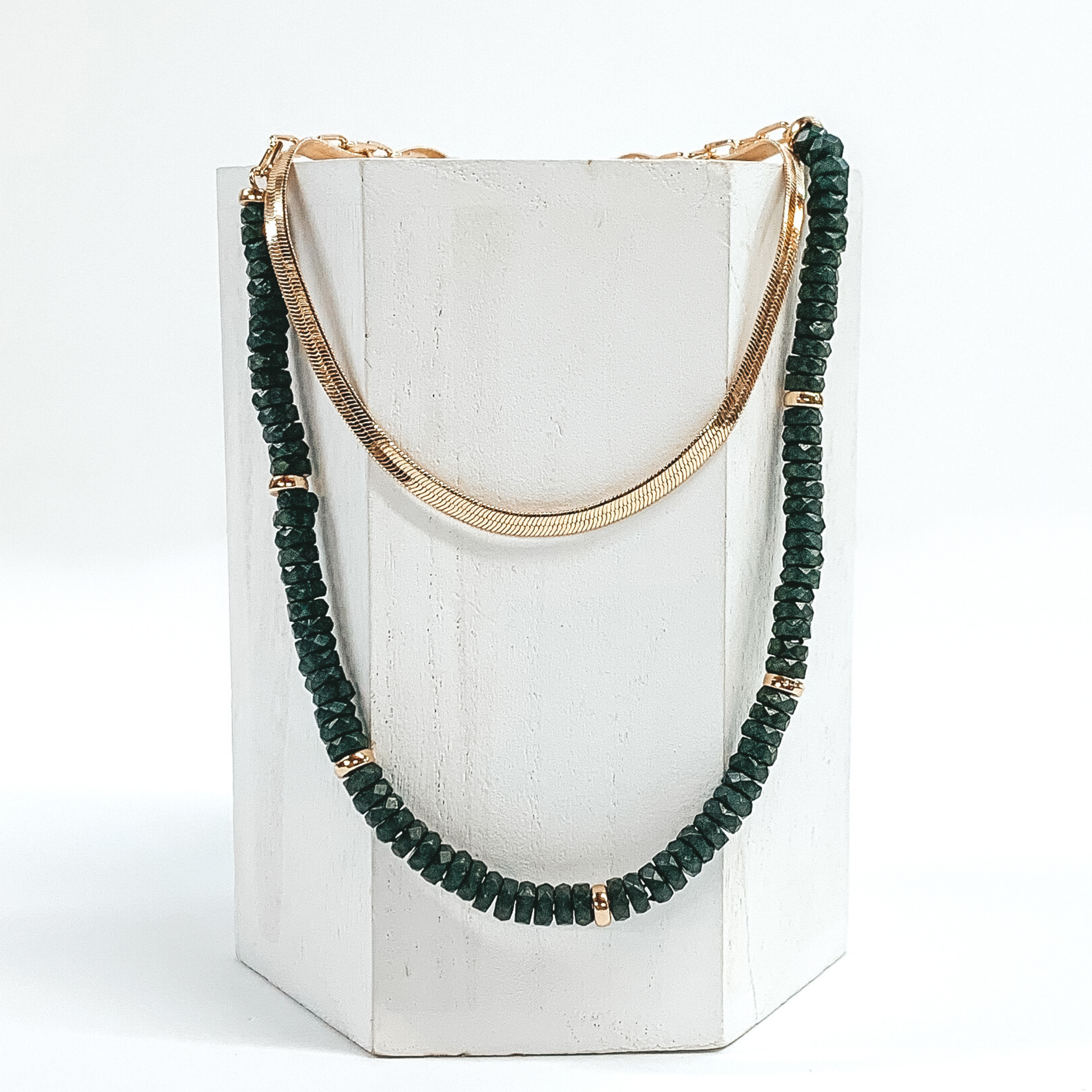 This necklace has a shorter gold snake chain and a longer olive green beaded necklace with gold bead spacers. This necklace is laid on a piece of white wood that is pictured on a white background. 