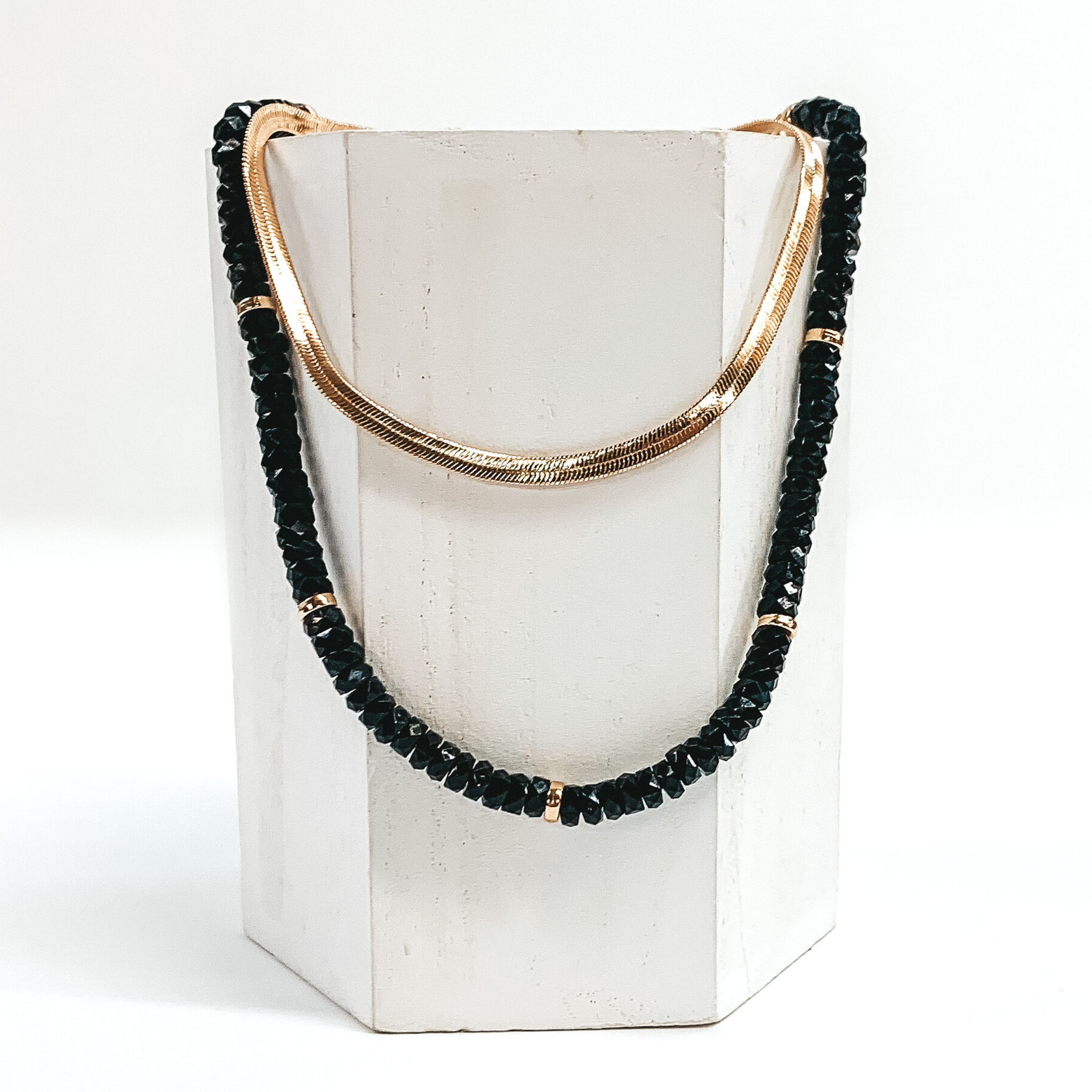This necklace has a shorter gold snake chain and a longer black beaded necklace with gold bead spacers. This necklace is laid on a piece of white wood that is pictured on a white background.
