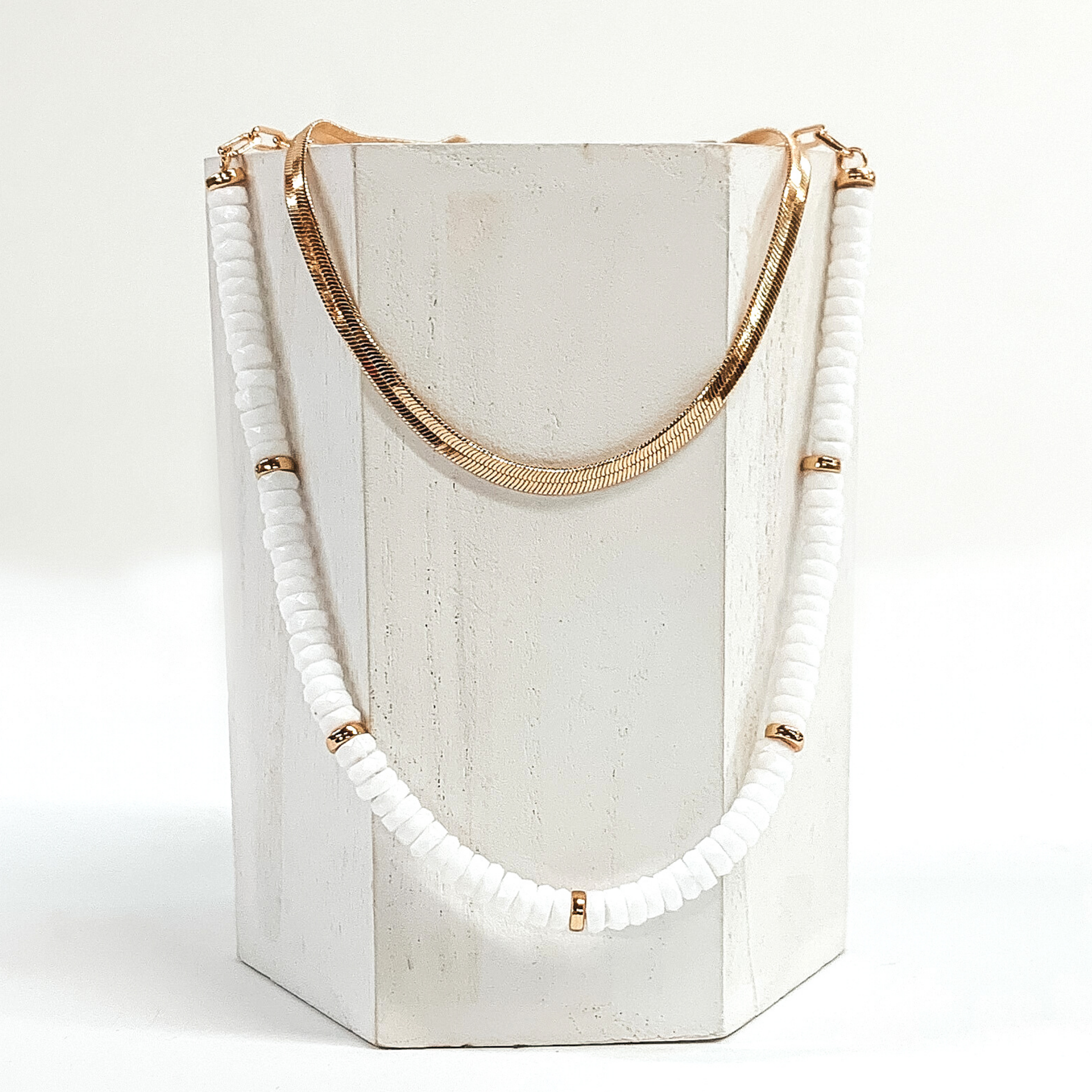 This necklace has a shorter gold snake chain and a longer white beaded necklace with gold bead spacers. This necklace is laid on a piece of white wood that is pictured on a white background.