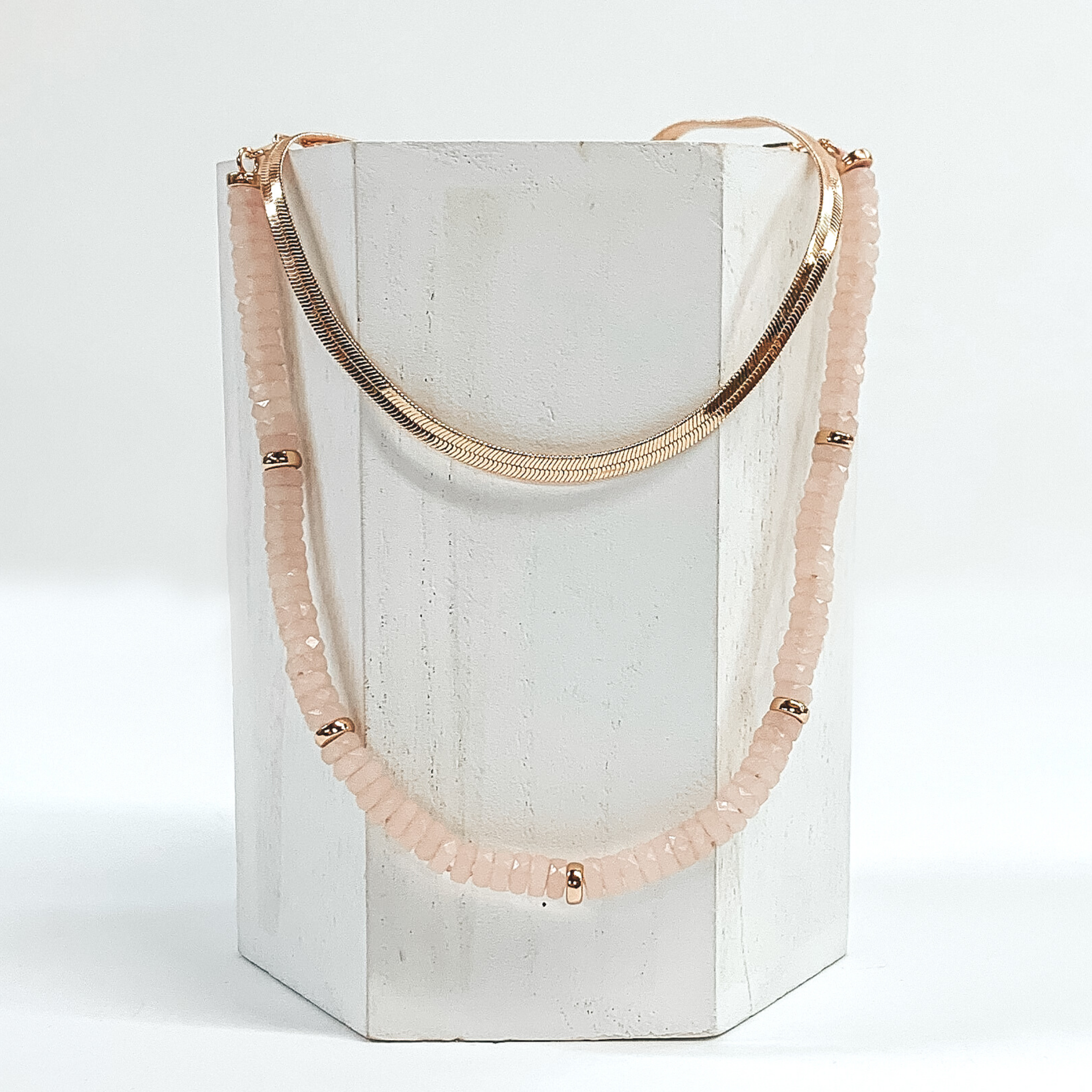 This necklace has a shorter gold snake chain and a longer blush beaded necklace with gold bead spacers. This necklace is laid on a piece of white wood that is pictured on a white background.