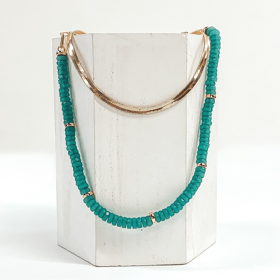 This necklace has a shorter gold snake chain and a longer teal beaded necklace with gold bead spacers. This necklace is laid on a piece of white wood that is pictured on a white background.