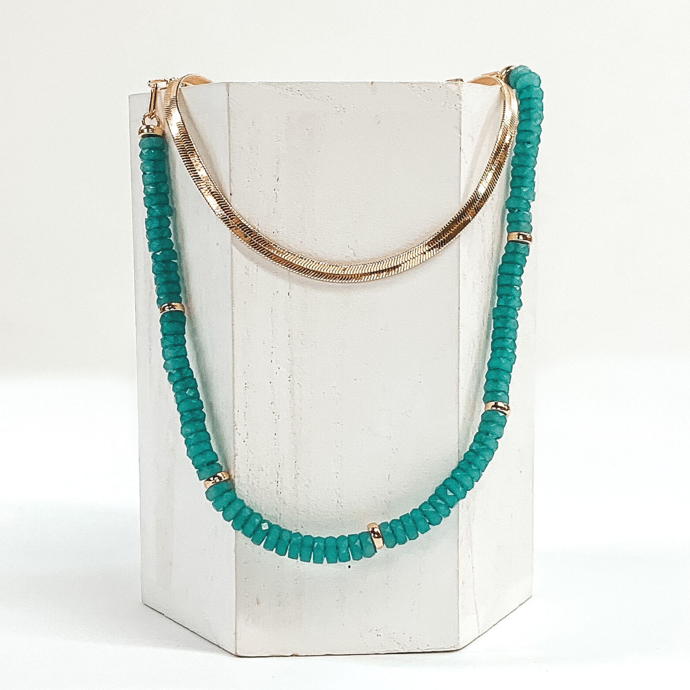 This necklace has a shorter gold snake chain and a longer teal beaded necklace with gold bead spacers. This necklace is laid on a piece of white wood that is pictured on a white background.