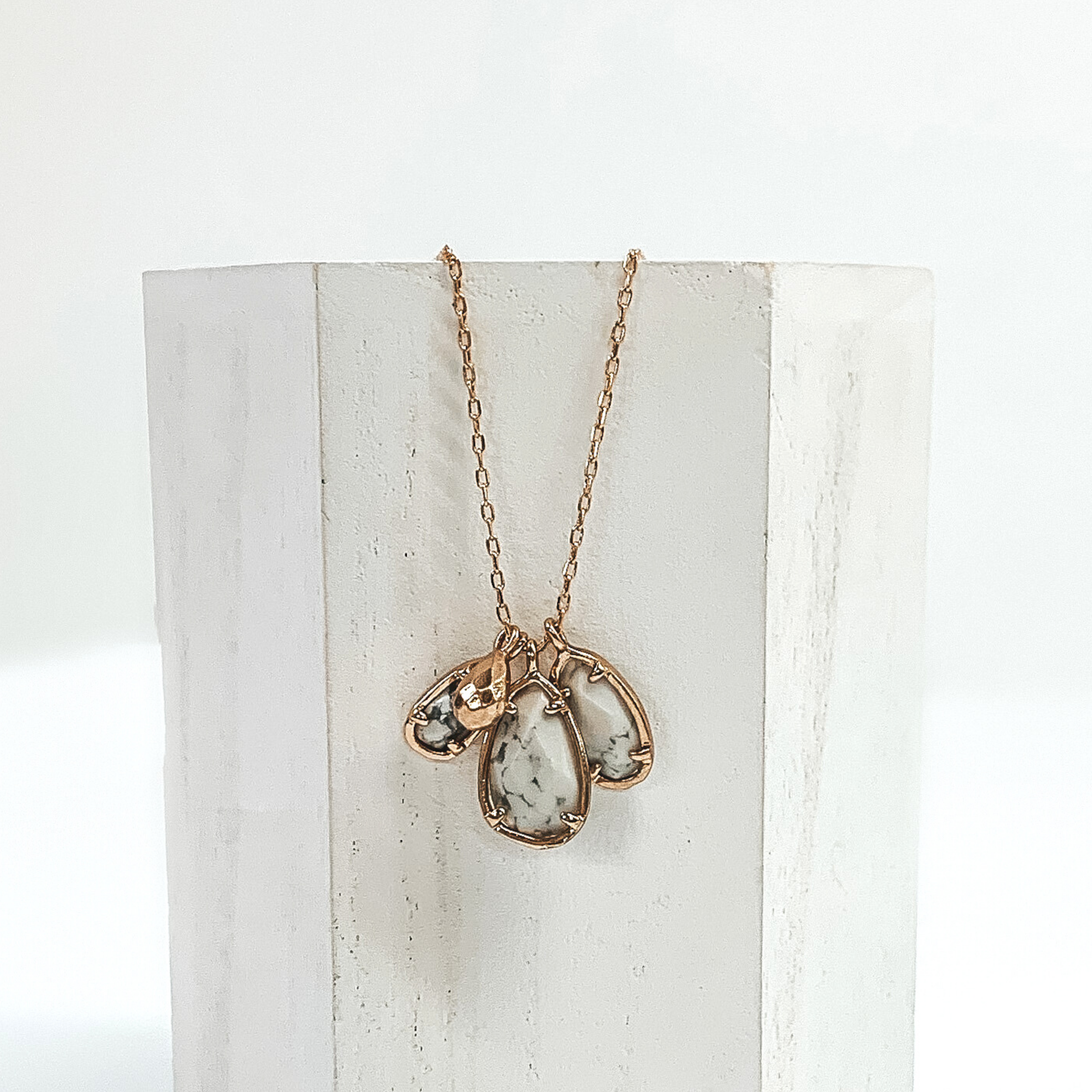 Small gold chain with three white marbled teardrop stone pendants outlined in gold. It also has a tiny gold teardrop pendant. This necklace is pictured laying a white piece of wood on a white background. 