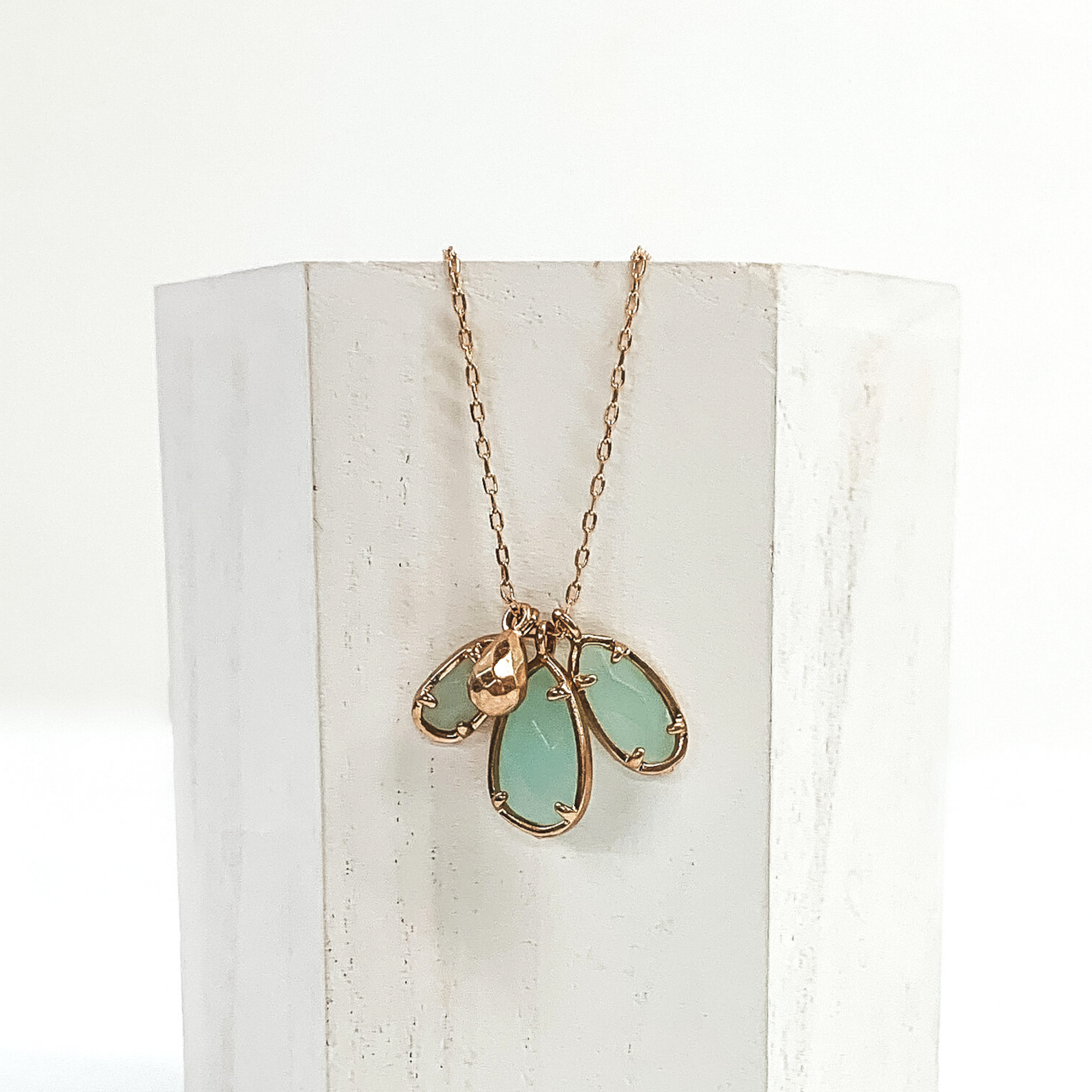 Small gold chain with three sage marbled teardrop stone pendants outlined in gold. It also has a tiny gold teardrop pendant. This necklace is pictured laying a white piece of wood on a white background.