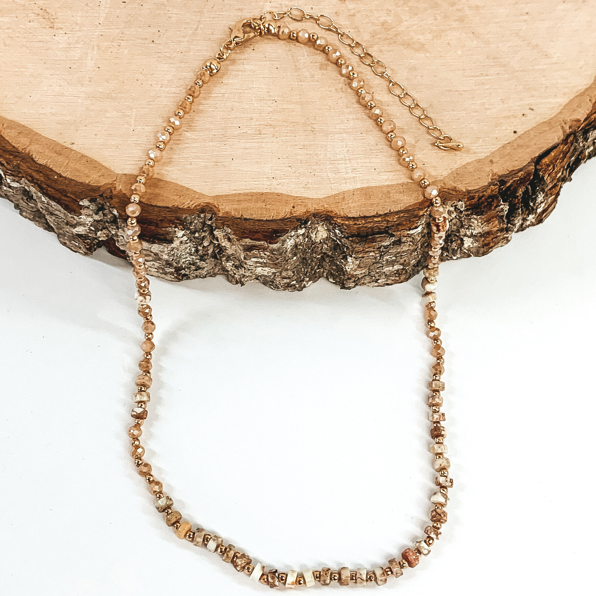 Tan marbled beads, tan crystal beads, and gold spacer beads strung together on an adjustable necklace. This necklace is pictured partly laying on a piece of wood on a white background. 