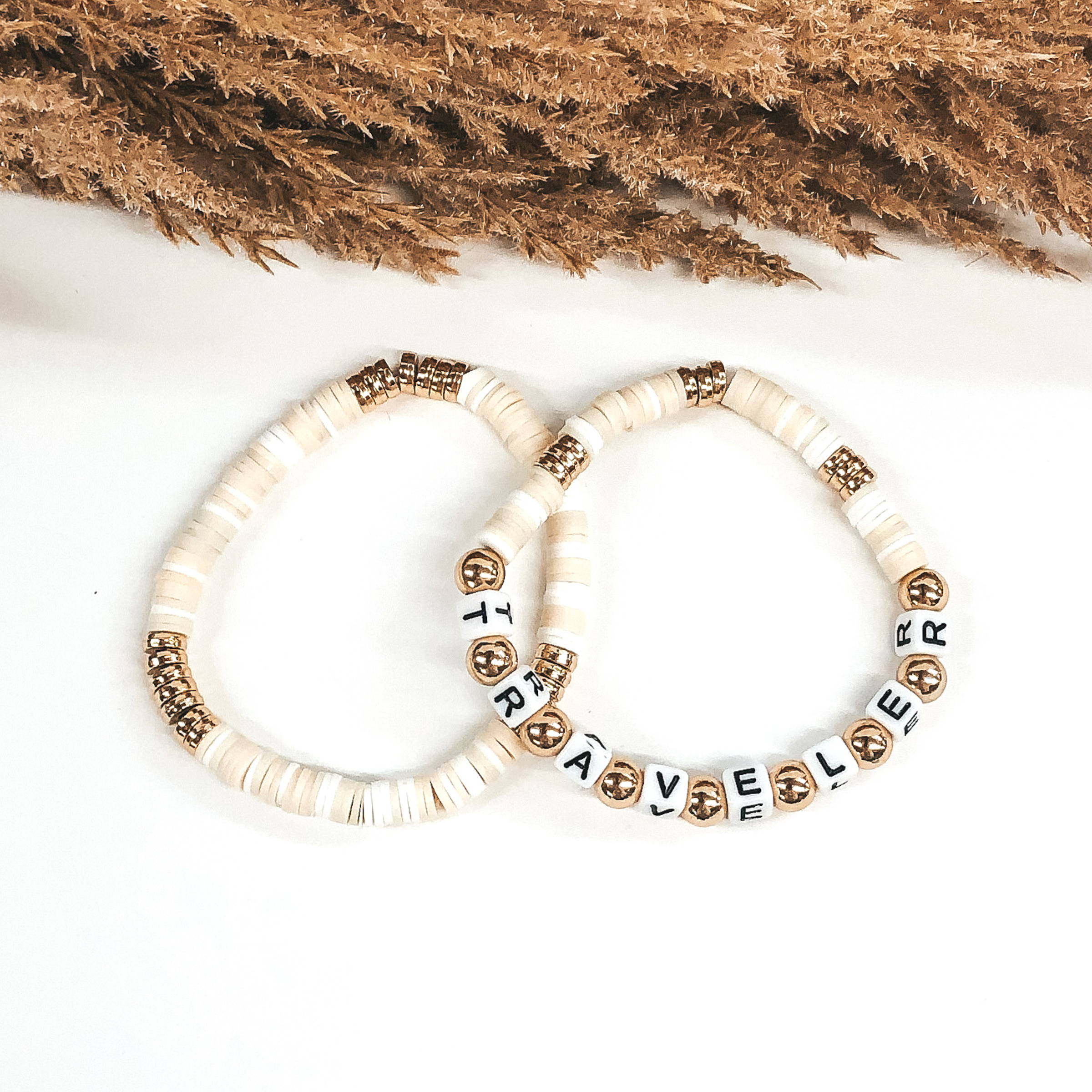 Two bracelets that has a mix of ivory colors and white disk beads. On one bracelet, you have three sections of gold disc beads. On the other bracelet you have three smaller section of gold disc beads and then letter beads that spell out "TRAVELER" with gold spacers in between. These bracelets are pictured on a white background.