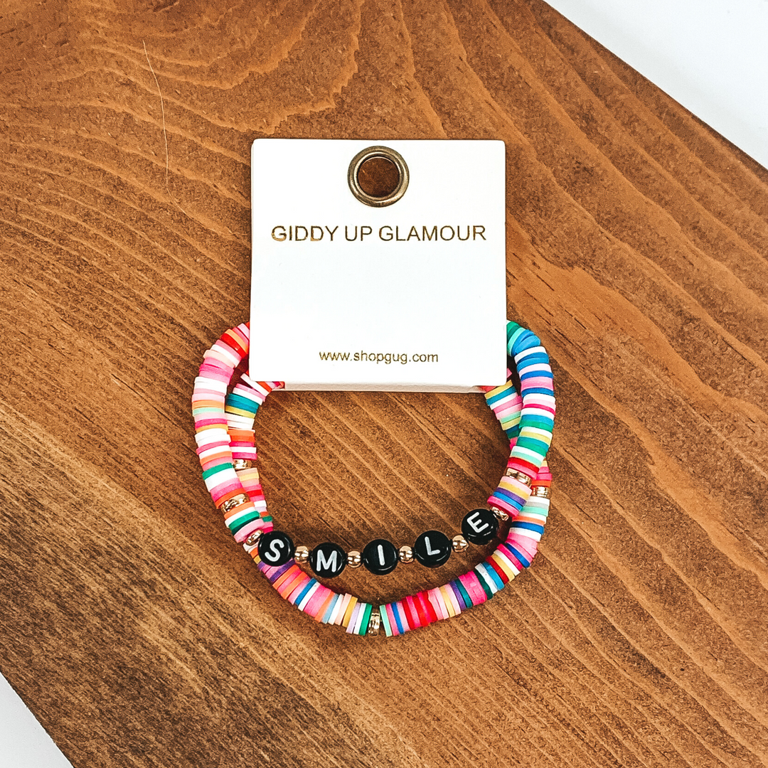Two bracelets that have multicolored disk beads. On one of the bracelets it has some gold spacers. On the other bracelet you have gold spacers and then black letter beads that spell out "SMILE" with gold spacers in between. These bracelets are pictured on a piece of wood on a white background.