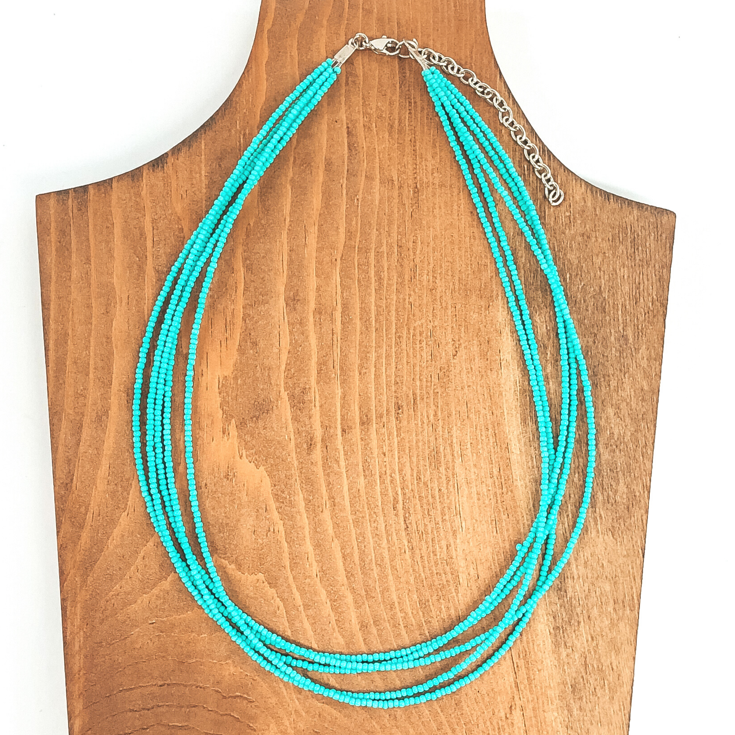 This necklace has multiple layered of seed beaded turquoise strands. This necklace is pictured laying on a wood necklace holder that is on a white background. 