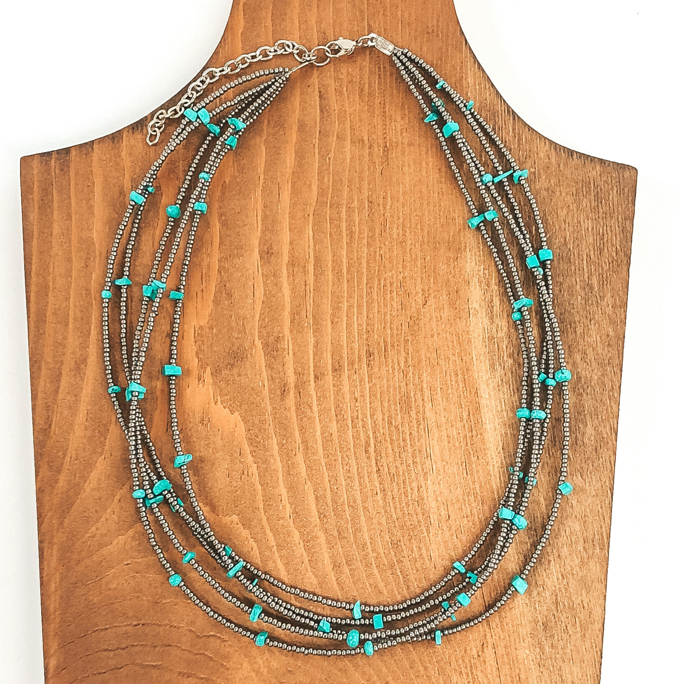 This necklace has multiple layers of seed beaded silver strands. Each strand also has turquoise colored stone spacers that haver irregular shapes. This necklace is pictured laying on a wood necklace holder that is on a white background.