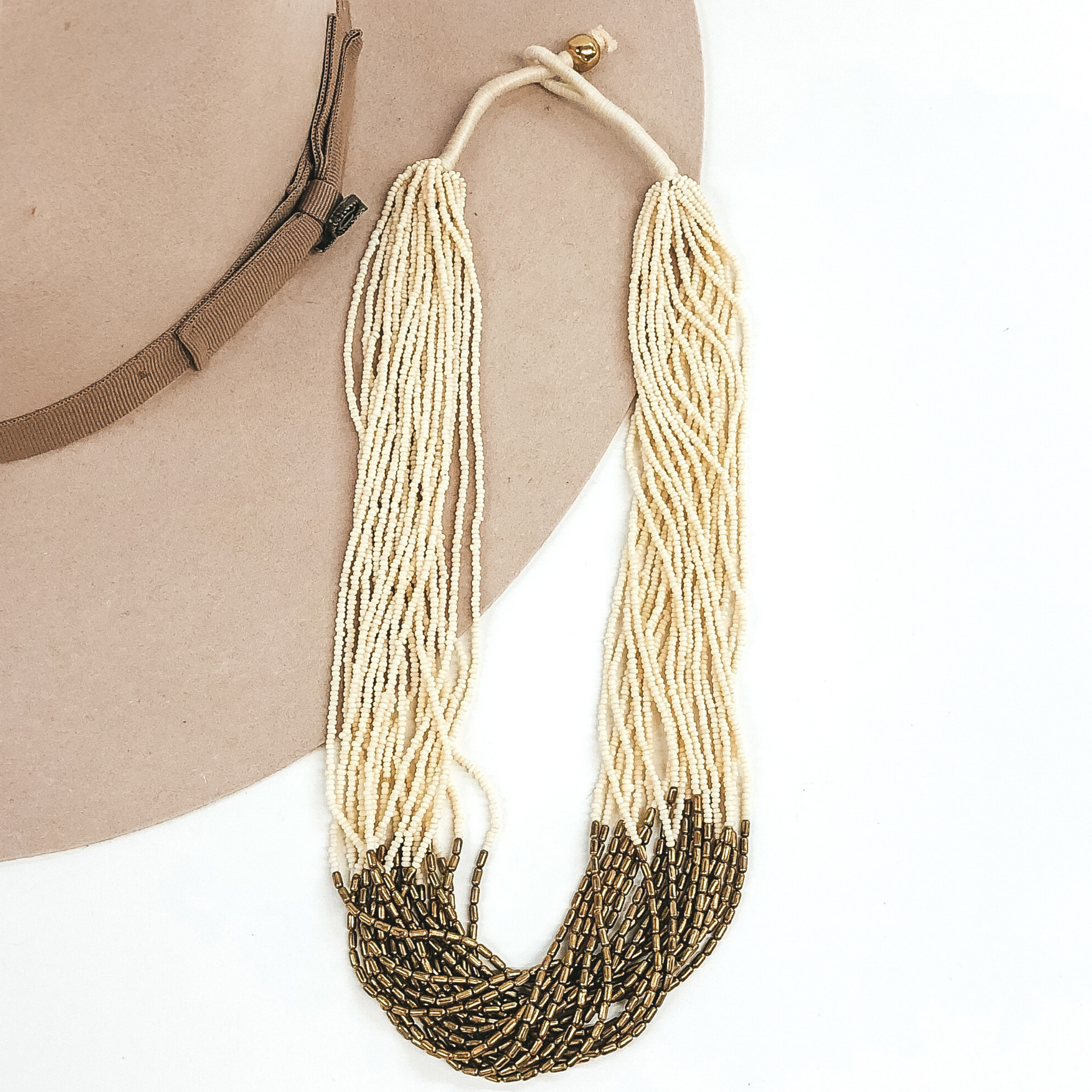The strands on this necklace have ivory beads and gold beads in the middle of the string. There are a lot of single strings put together to make a single necklace. The necklace has a gold bead that goes through a loop to put the necklace on. This necklace is pictured laying on a beige hat on a white background. 