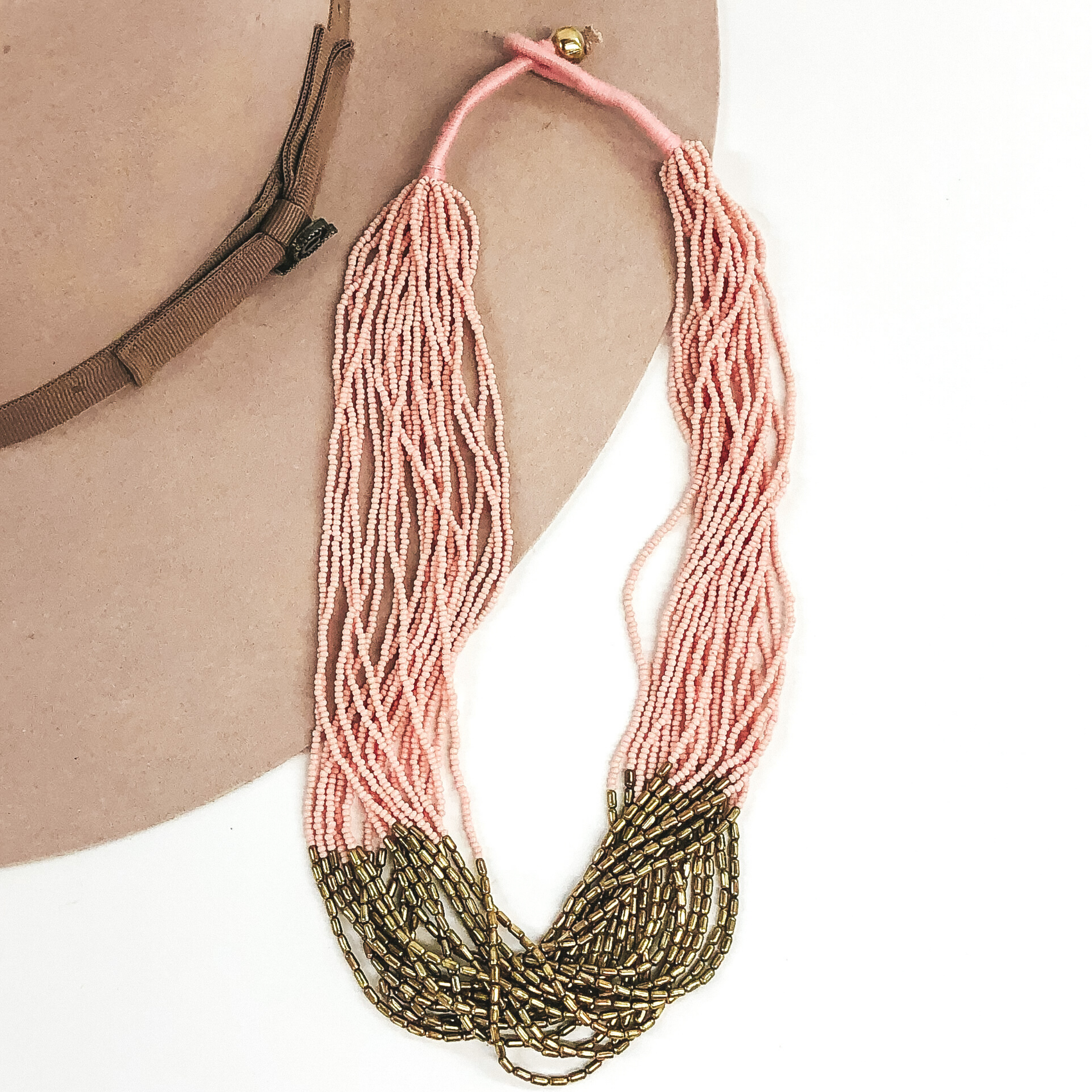 The strands on this necklace have light pink beads and gold beads in the middle of the string. There are a lot of single strings put together to make a single necklace. The necklace has a gold bead that goes through a loop to put the necklace on. This necklace is pictured laying on a beige hat on a white background.