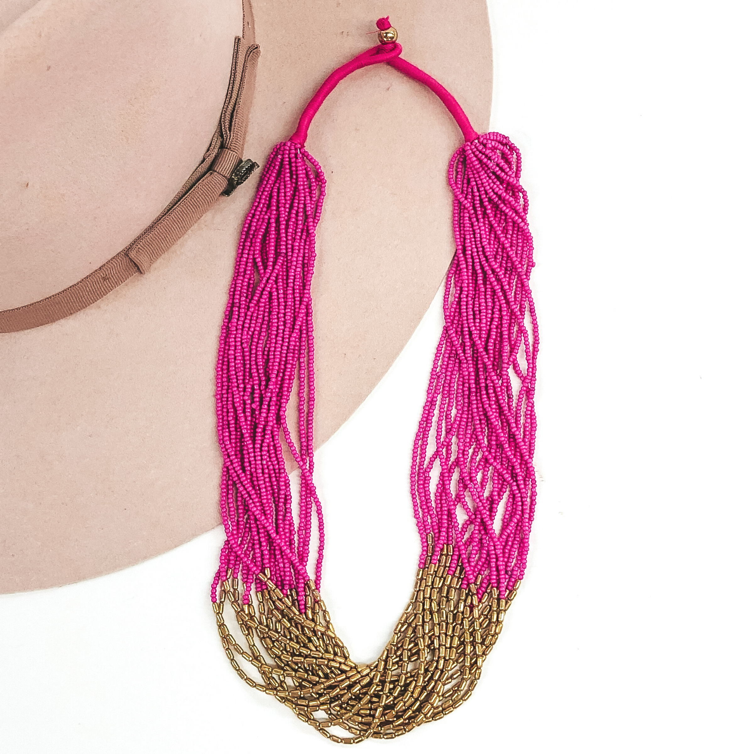 The strands on this necklace have fuchsia beads and gold beads in the middle of the string. There are a lot of single strings put together to make a single necklace. The necklace has a gold bead that goes through a loop to put the necklace on. This necklace is pictured laying on a beige hat on a white background.