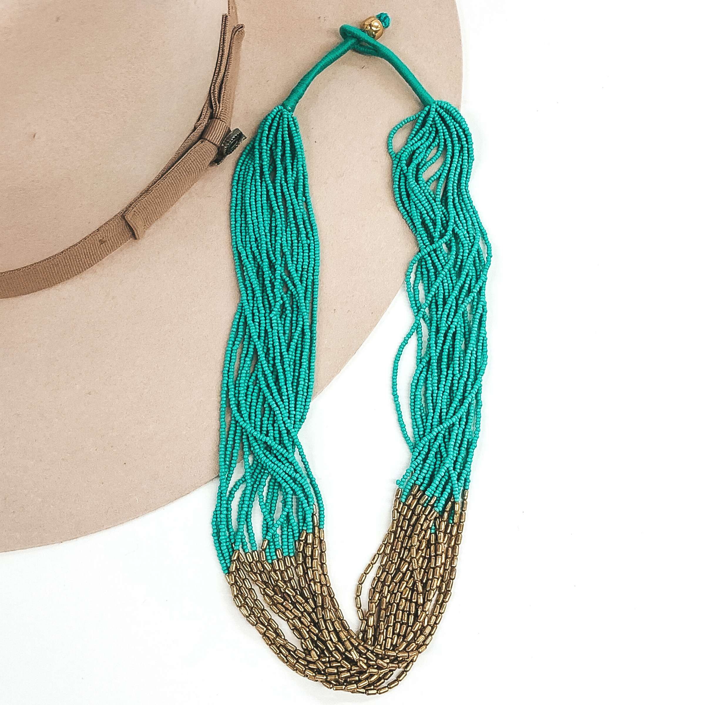 The strands on this necklace have turquoise beads and gold beads in the middle of the string. There are a lot of single strings put together to make a single necklace. The necklace has a gold bead that goes through a loop to put the necklace on. This necklace is pictured laying on a beige hat on a white background.