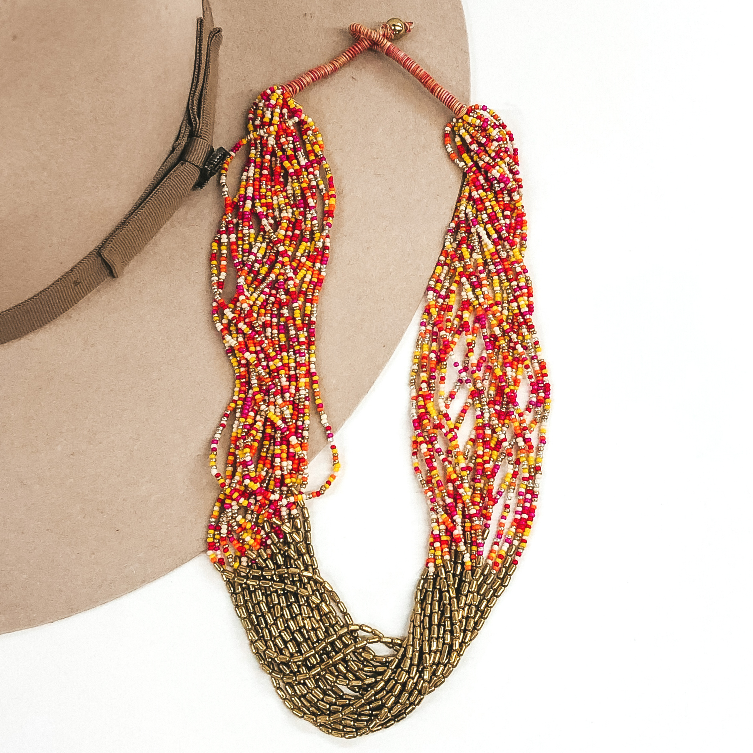 The strands on this necklace have multicolored beads and gold beads in the middle of the string. There are a lot of single strings put together to make a single necklace. The necklace has a gold bead that goes through a loop to put the necklace on. This necklace is pictured laying on a beige hat on a white background.