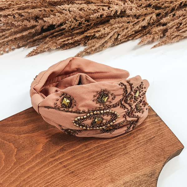 This is a thick, dusty mauve colored headband with gold and bronze beaded detailing. The headband also comes together in the middle with a piece of fabric keeping it small and tied together. This headband is pictured laying in a piece of wood with tan floral at the top of the picture on a white background.