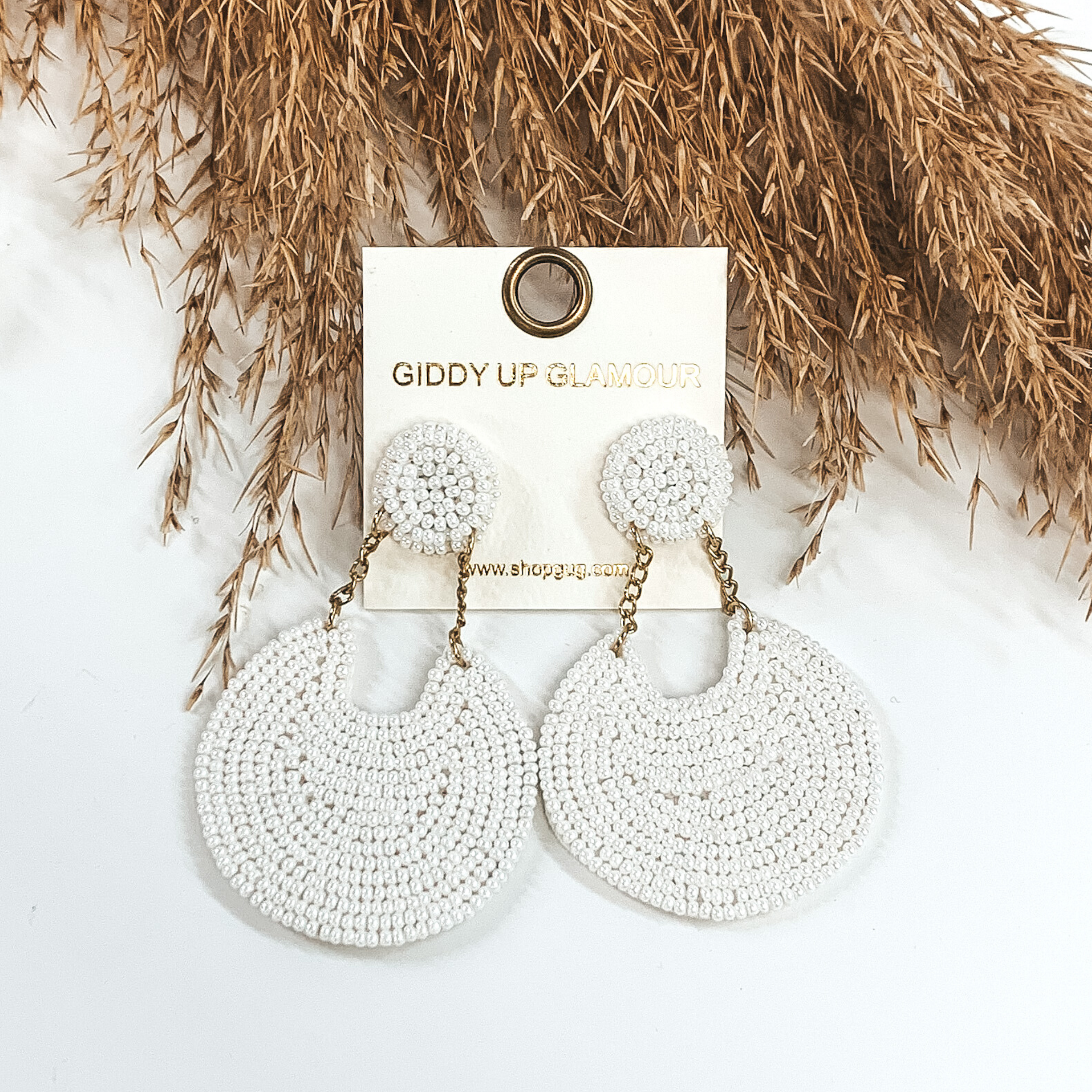 These earrings are covered in white beads. There is a circle stud with two gold chains that hold a bigger, hanging circle that has a notch out of the top. These earrings are pictured on a white background with tan floral at the top of the picture.