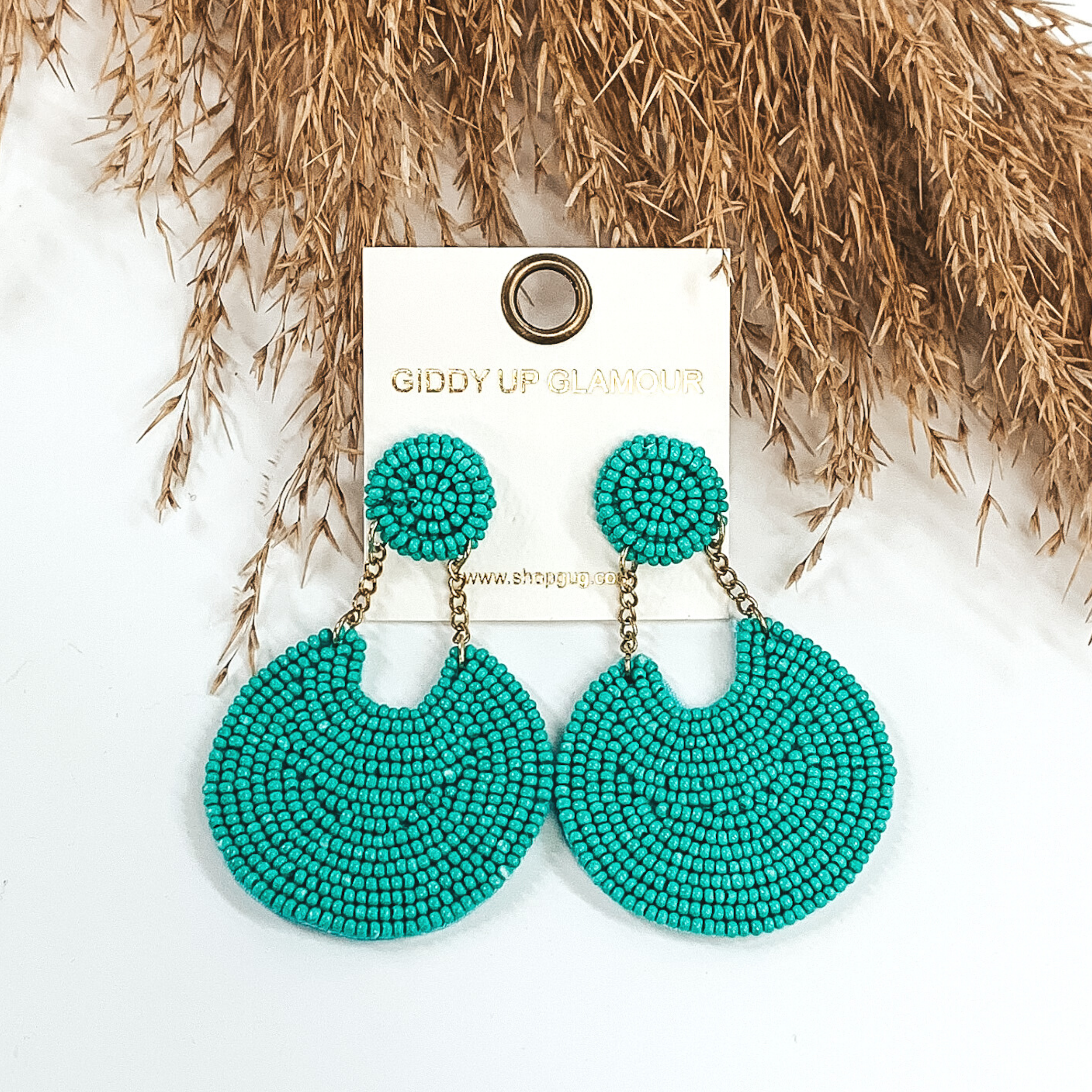 These earrings are covered in turquoise beads. There is a circle stud with two gold chains that hold a bigger, hanging circle that has a notch out of the top. These earrings are pictured on a white background with tan floral at the top of the picture.