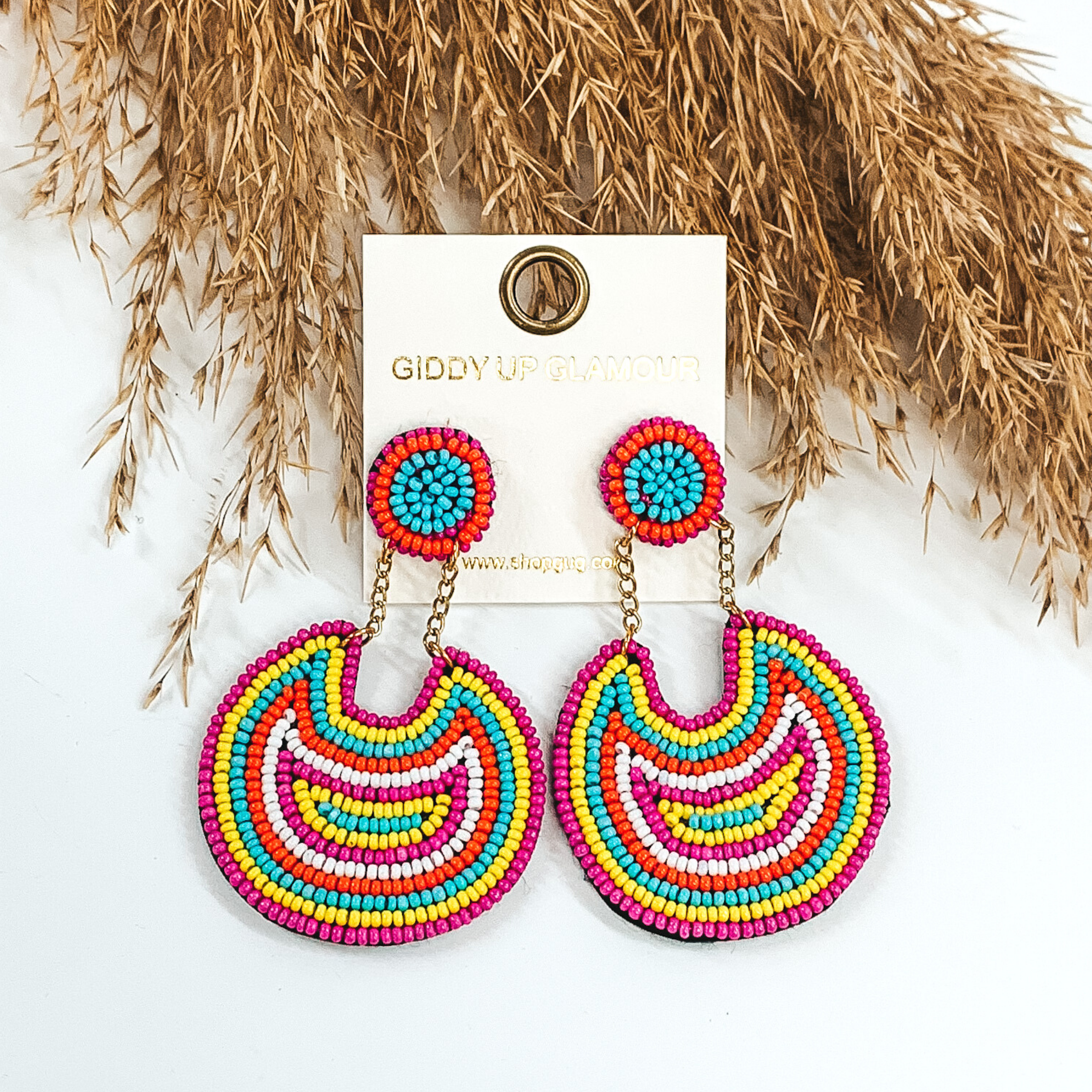 These earrings are covered in white, orange, blue, yellow, and fuchsia beads. There is a circle stud with two gold chains that hold a bigger, hanging circle that has a notch out of the top. These earrings are pictured on a white background with tan floral at the top of the picture.