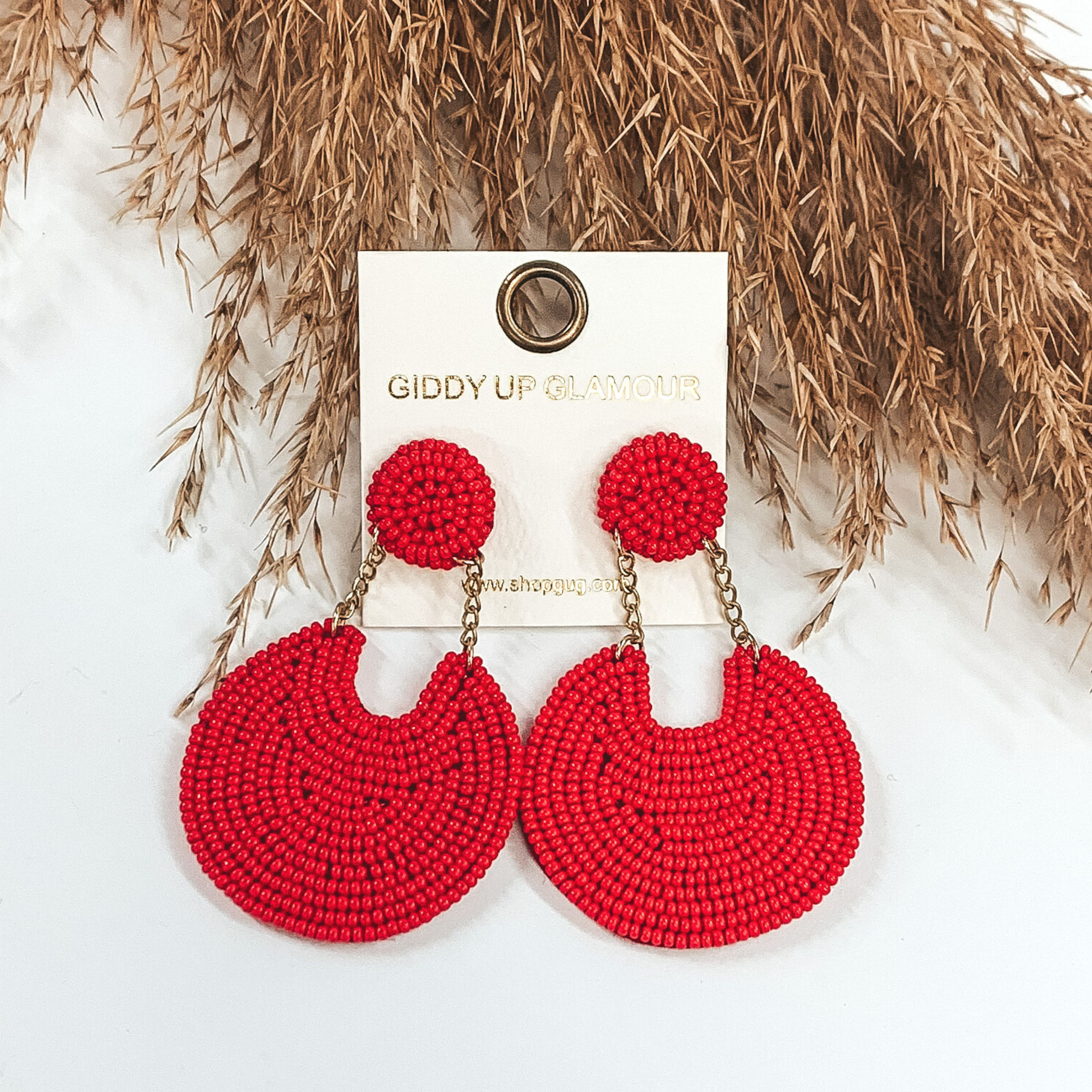 These earrings are covered in red beads. There is a circle stud with two gold chains that hold a bigger, hanging circle that has a notch out of the top. These earrings are pictured on a white background with tan floral at the top of the picture.
