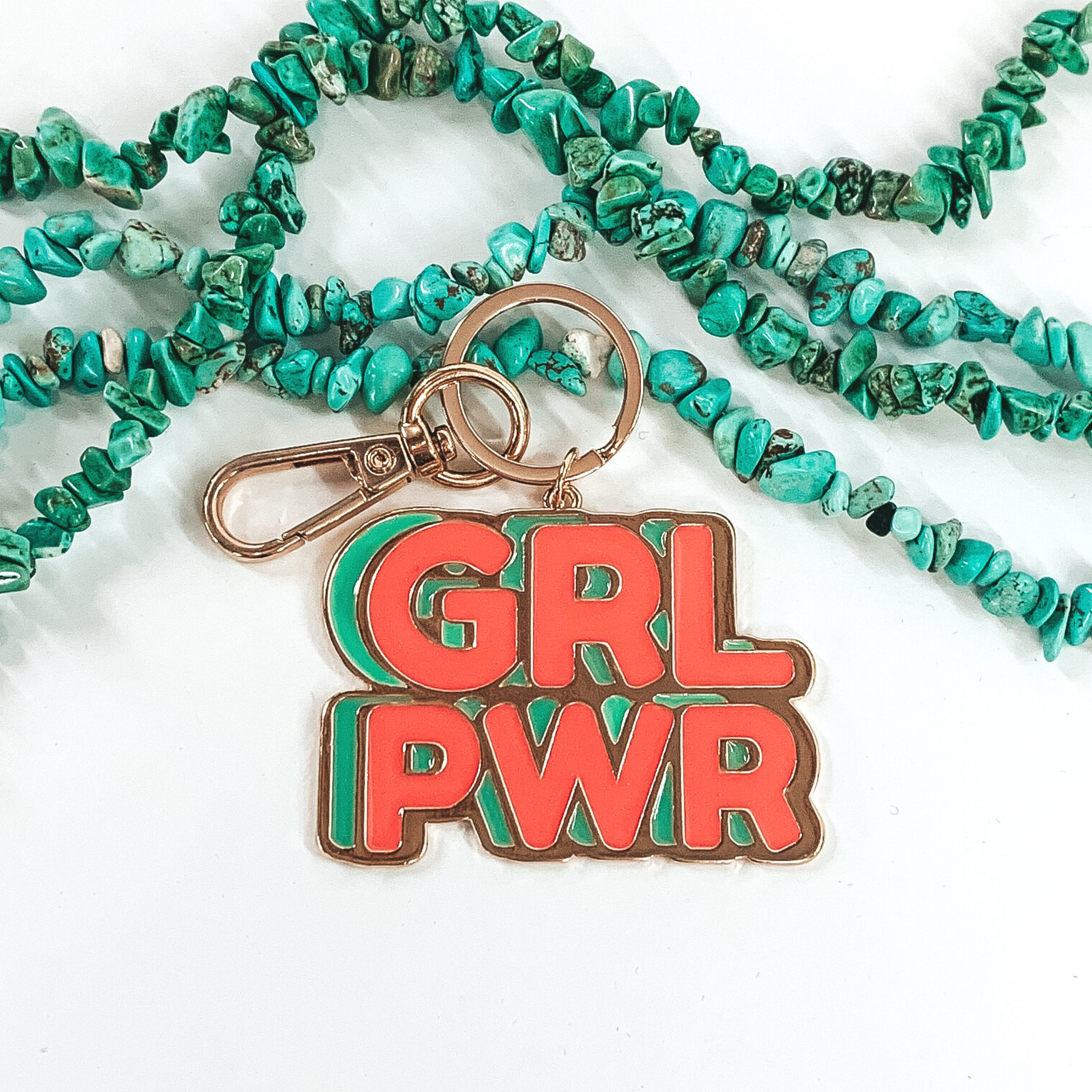 This is a gold key chain with a pendant that spells out "GRL PWR." The letters are in a coral color and is shadowed by a teal color. It is pictured on a white background with turquoise beads in at the top of the picture. 
