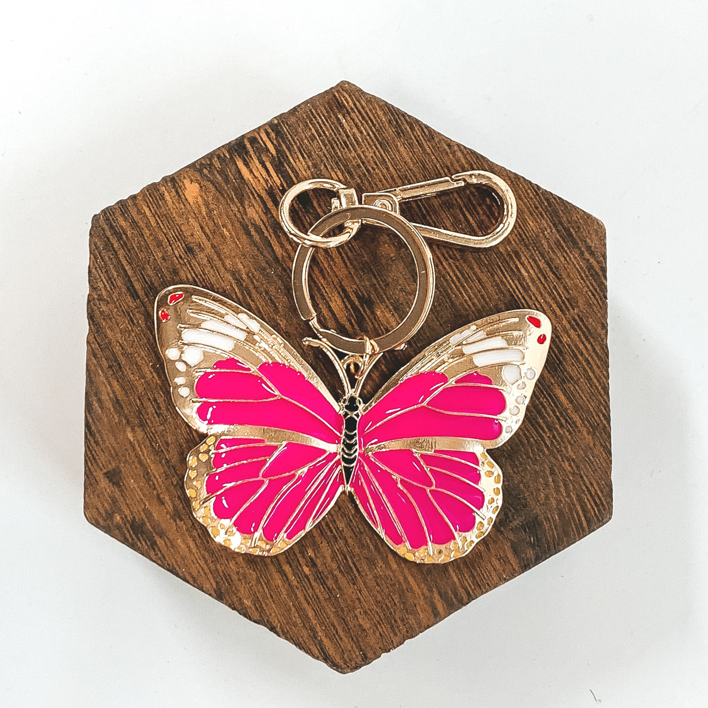 This is a gold key chain and a gold butterfly pendant that includes pink, white, and a hint of red color. It is pictured on a white background  laying on a dark brown hexagon. 