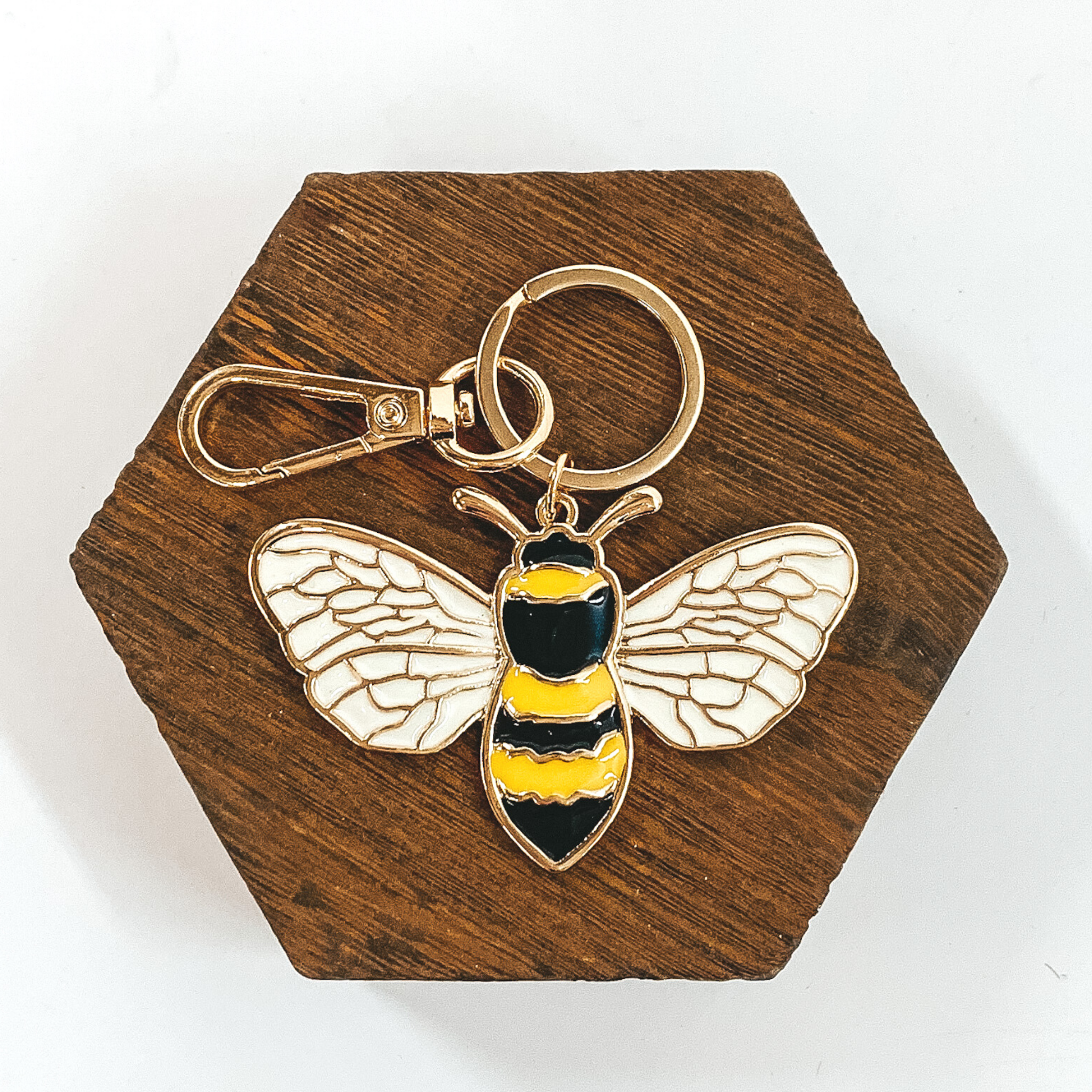 This is a gold key chain with a bumble bee pendant. The pendant includes white, yellow, and block colors. It is pictured on a white background laying on a dark brown hexagon.