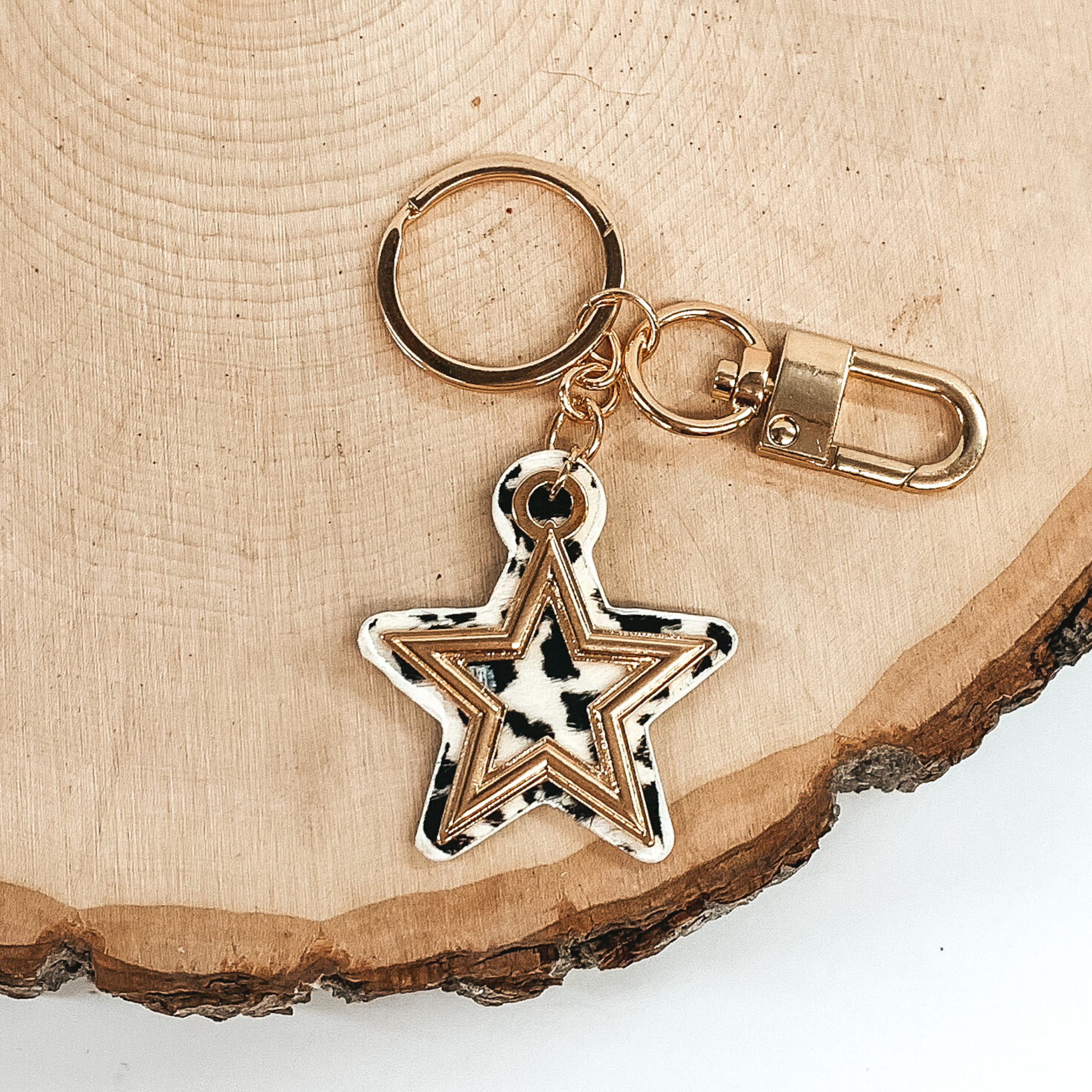 Gold key chain with a white and black leopard print star pendant that includes a gold star outline. This key chain is pictured on a piece of wood on a white background. 