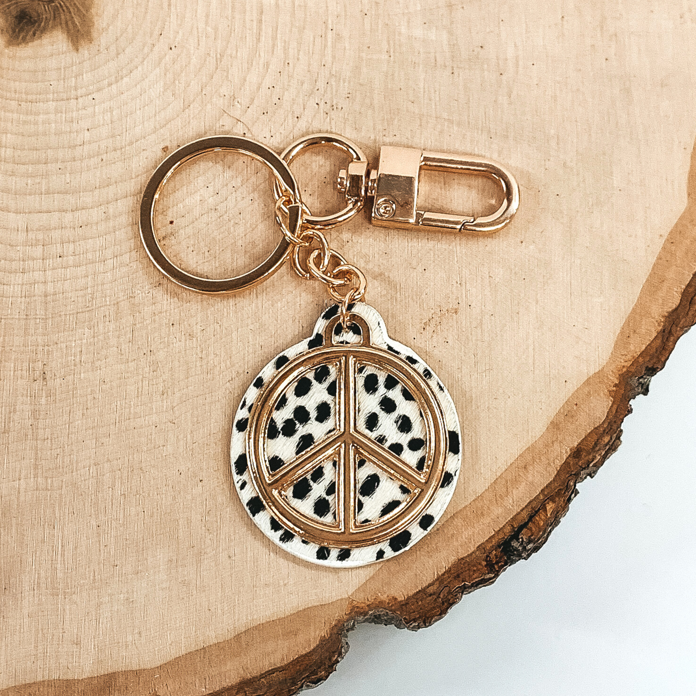 Gold key chain with a white and black dotted print circle pendant that includes a gold peace sign outline. This key chain is pictured on a piece of wood on a white background.