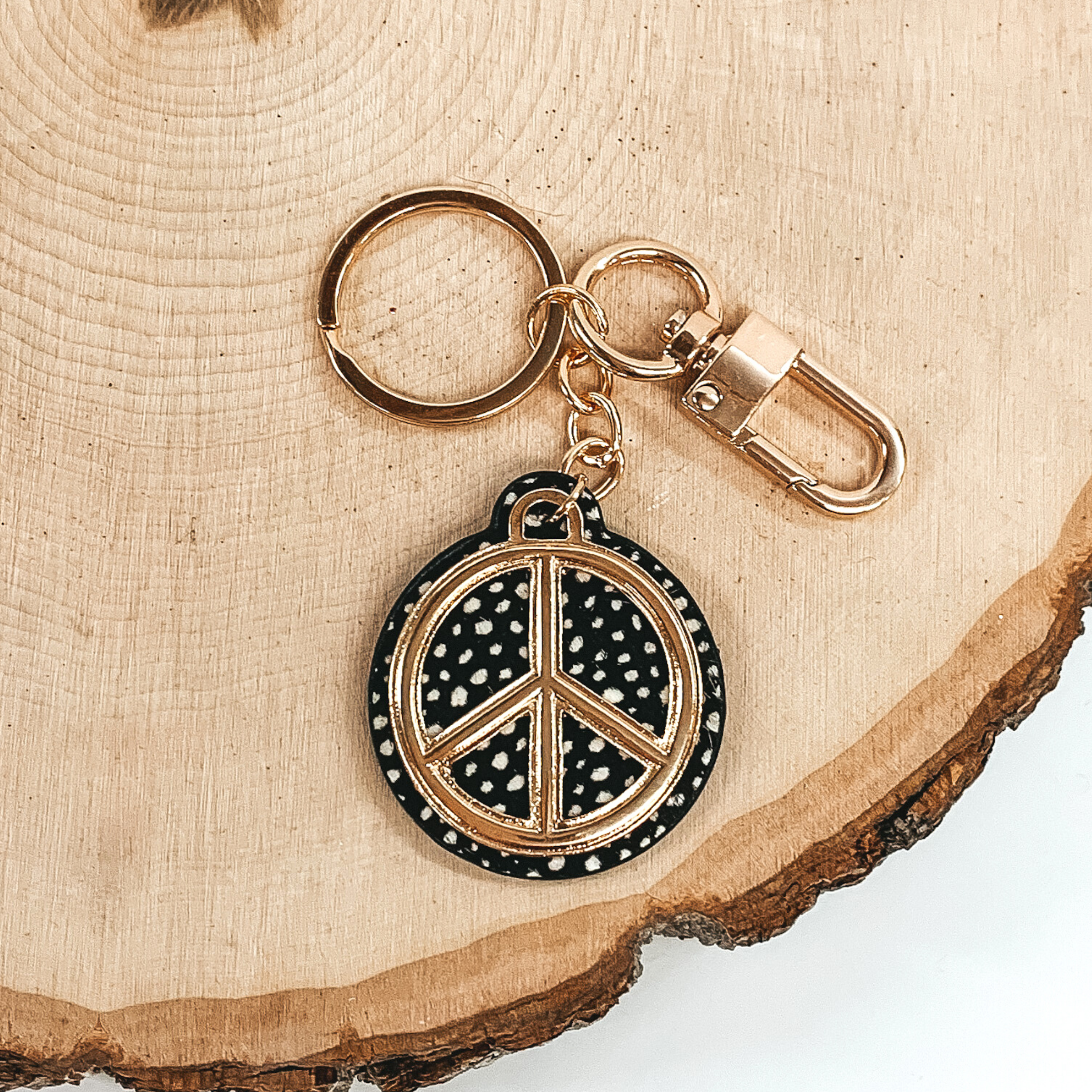 Gold key chain with a black and white dotted print circle pendant that includes a gold peace sign outline. This key chain is pictured on a piece of wood on a white background.