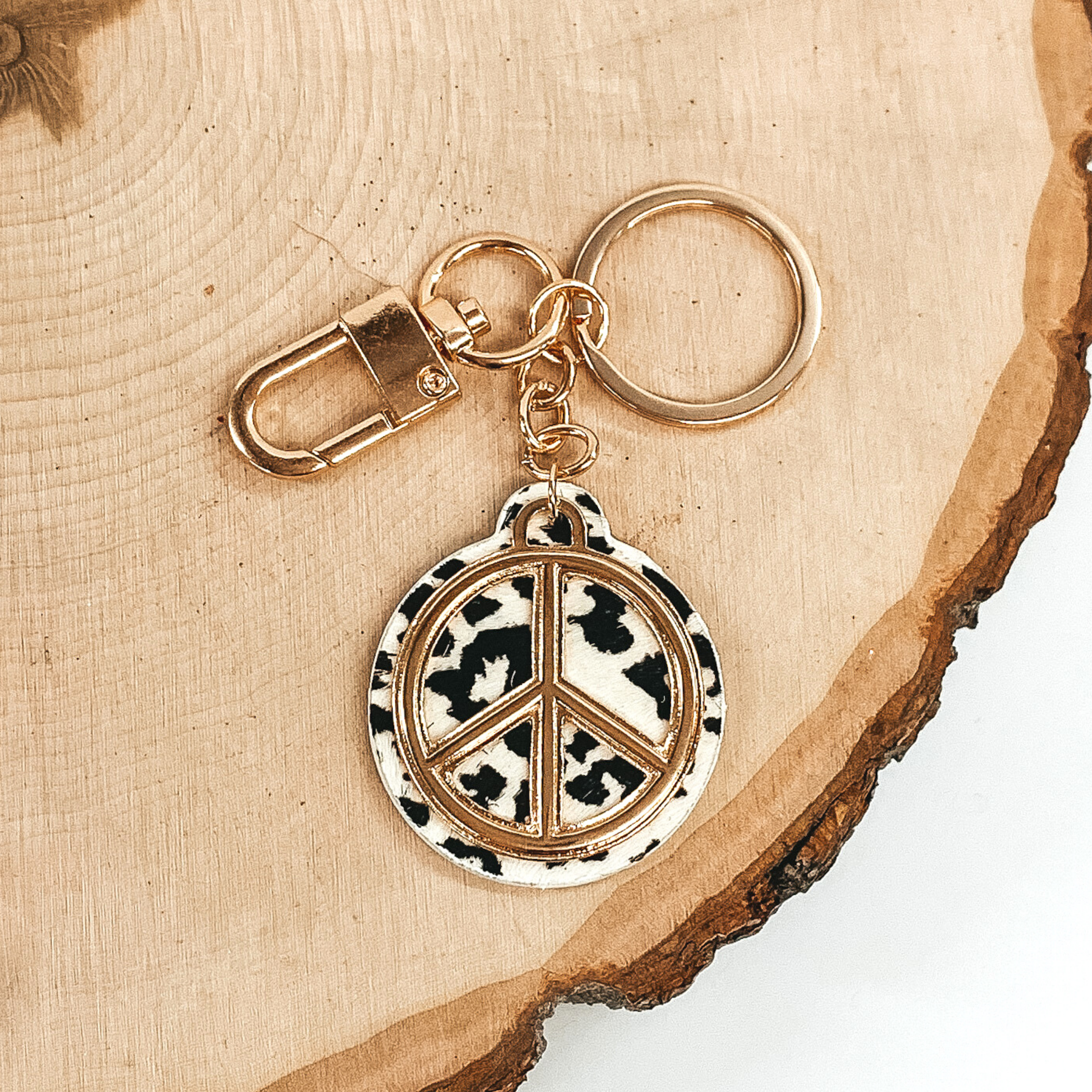 Gold key chain with a white and black leopard print circle pendant that includes a gold peace sign outline. This key chain is pictured on a piece of wood on a white background.
